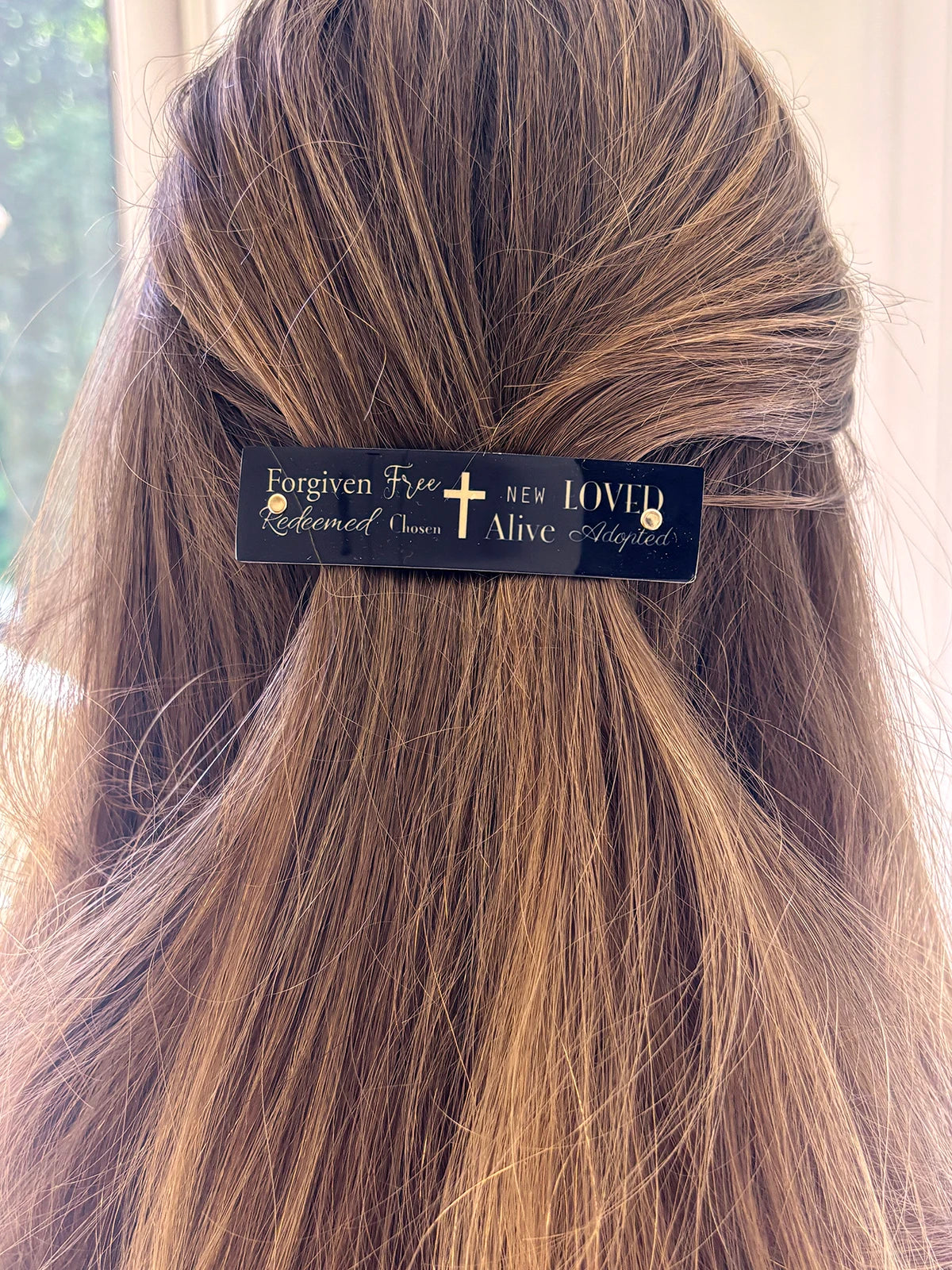 A close-up of a person's brown hair secured with a black hair clip featuring gold religious text that reads 'Forgiven Free  Redeemed Chosen  New Loved Alive Adopted'.