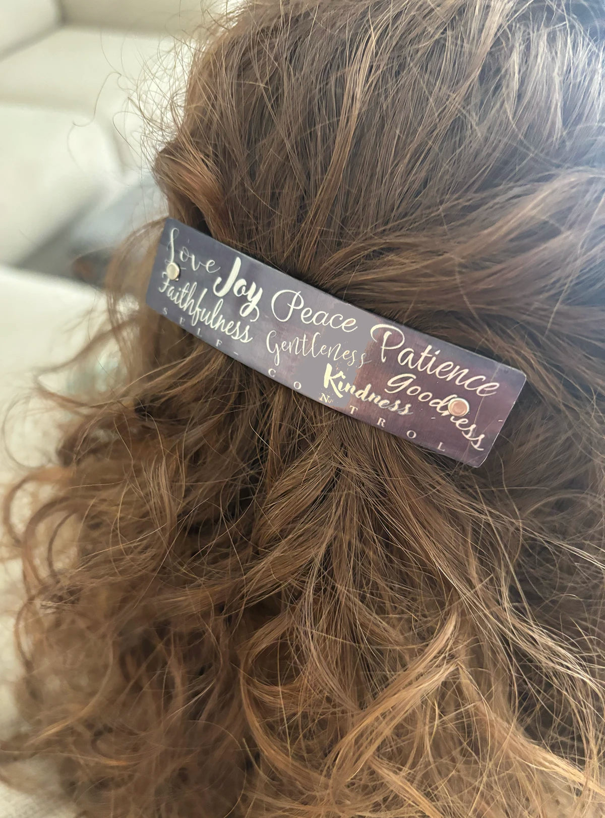 A close-up of brown wavy hair secured with a dark rectangular hair clip featuring silver script words including 'Love', 'Joy', 'Peace', 'Patience', 'Kindness', and 'Gentleness'. Handmade in North Carolina.