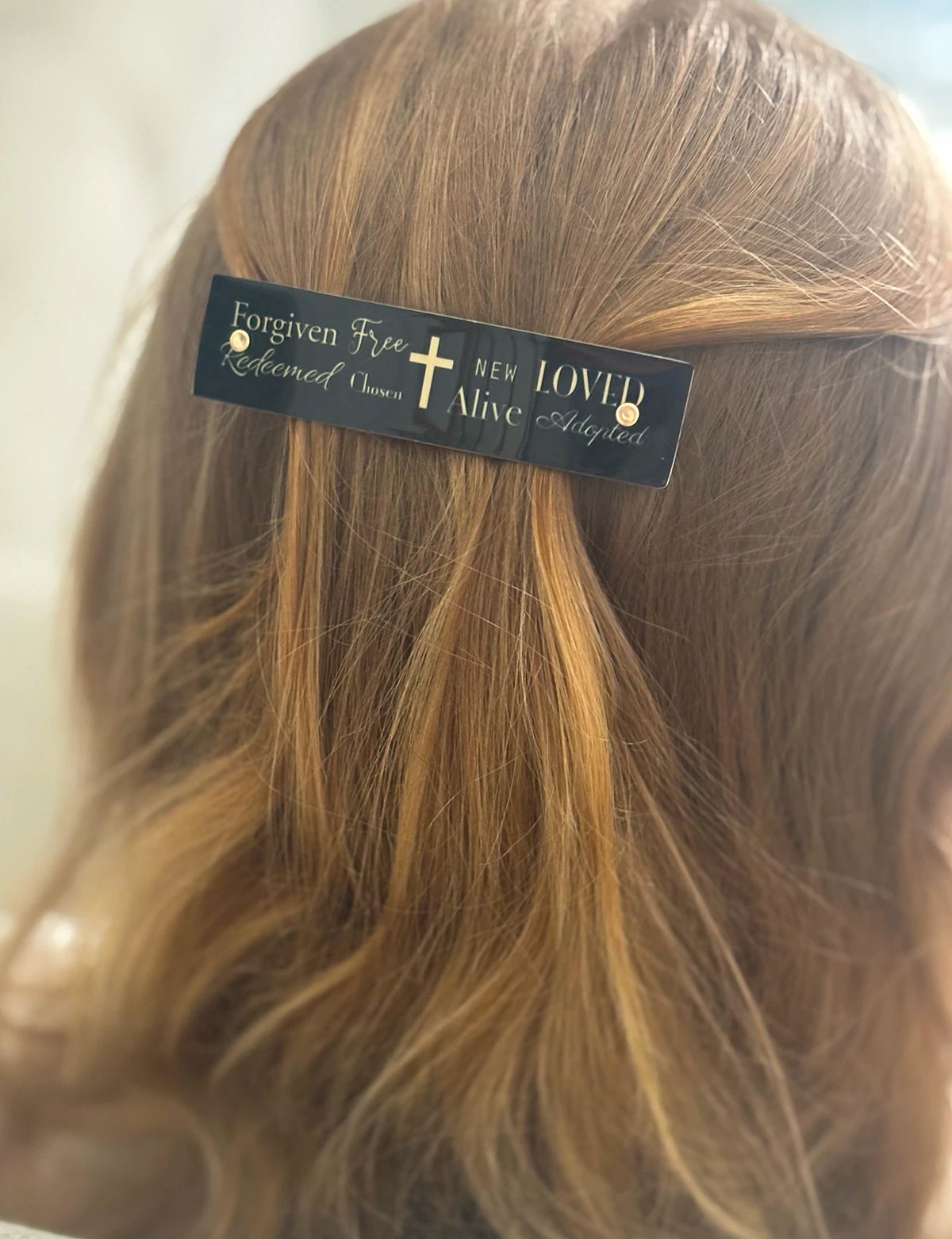 A close-up of a person's light brown hair with a black hair clip holding a section of hair. The clip has gold text reading 'Forgiven Free Redeemed Chose NEW LOVED Alive Adopted' with a gold cross in the center.