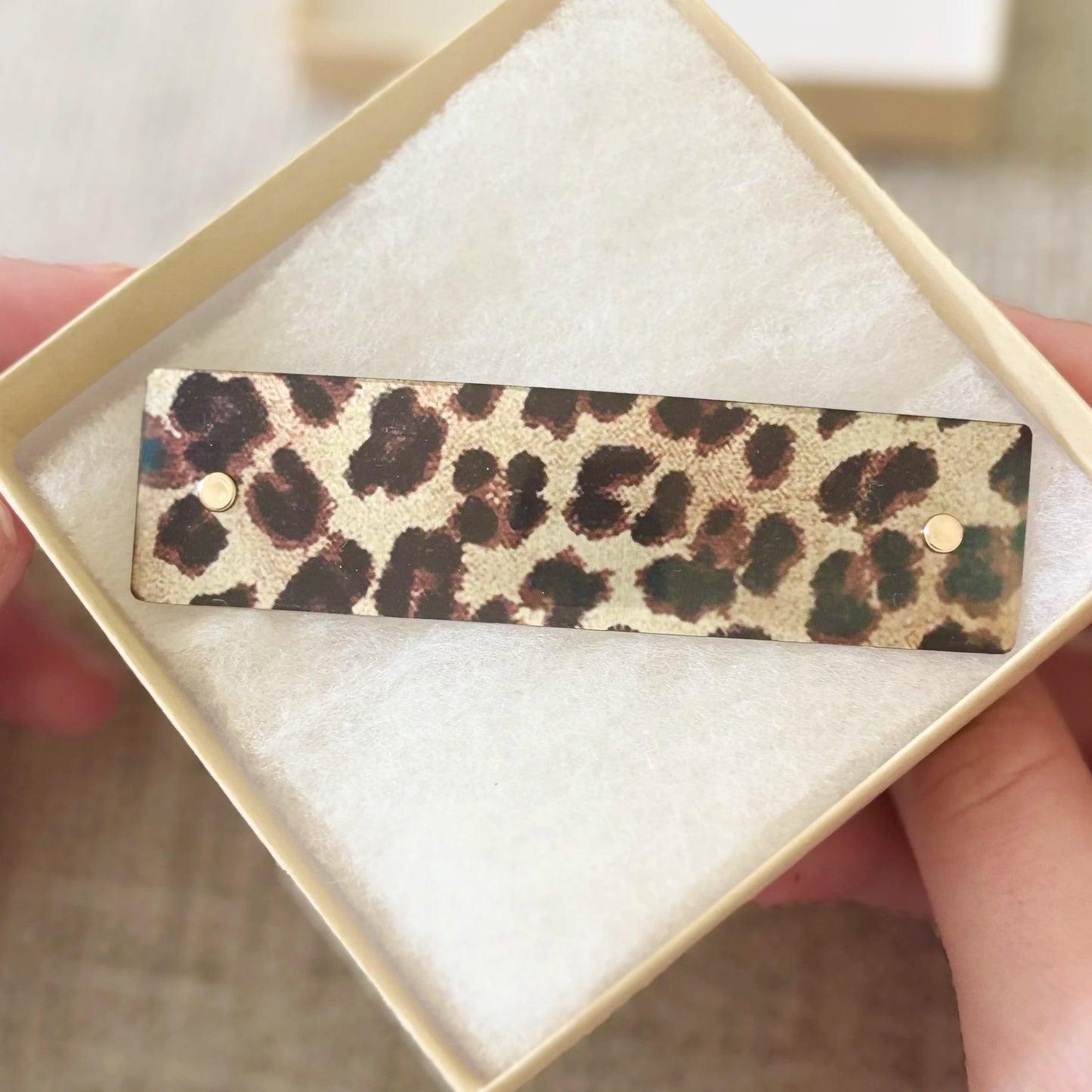 A leopard print barrette hair clip with gold-tone metal studs, presented inside a cream-colored square gift box with white padding, held by hands.