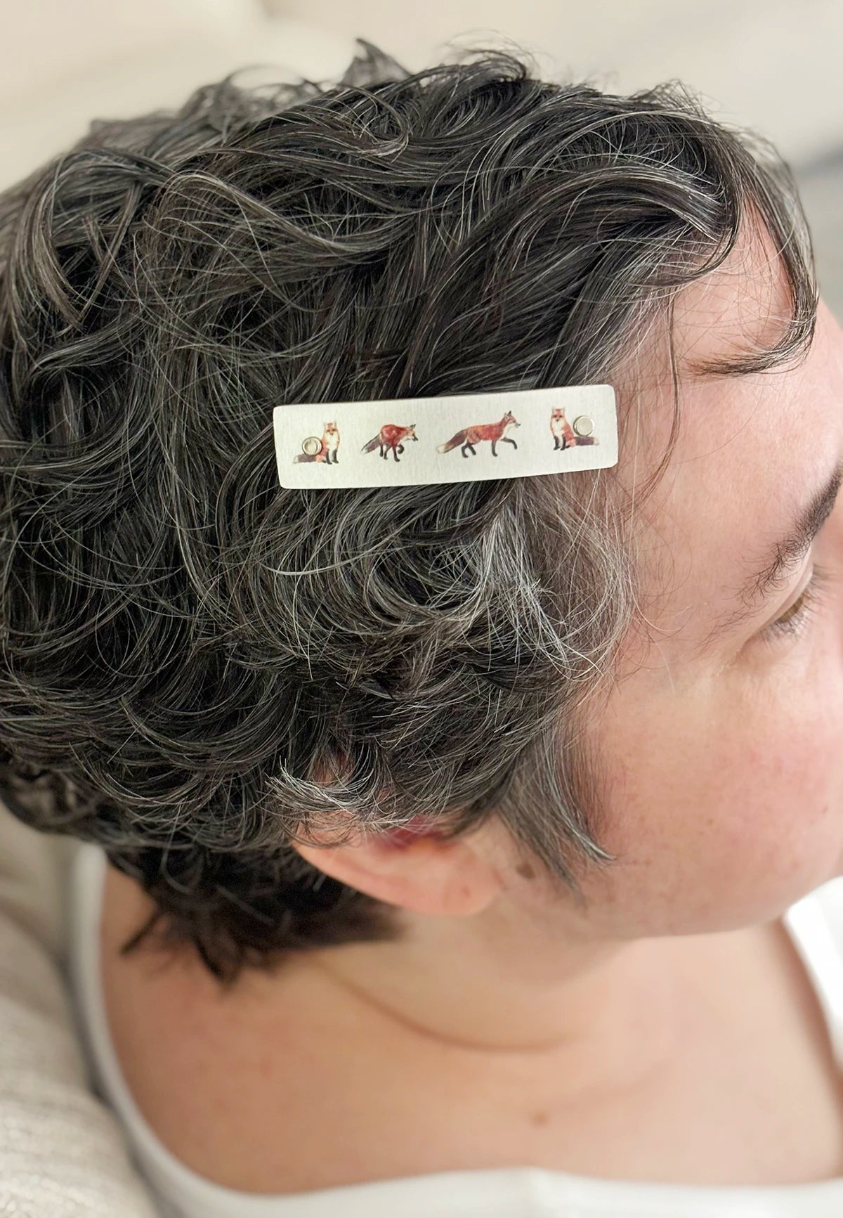A close-up of a person's short, curly gray hair wearing a silver hair clip decorated with small detailed illustrations of red foxes.