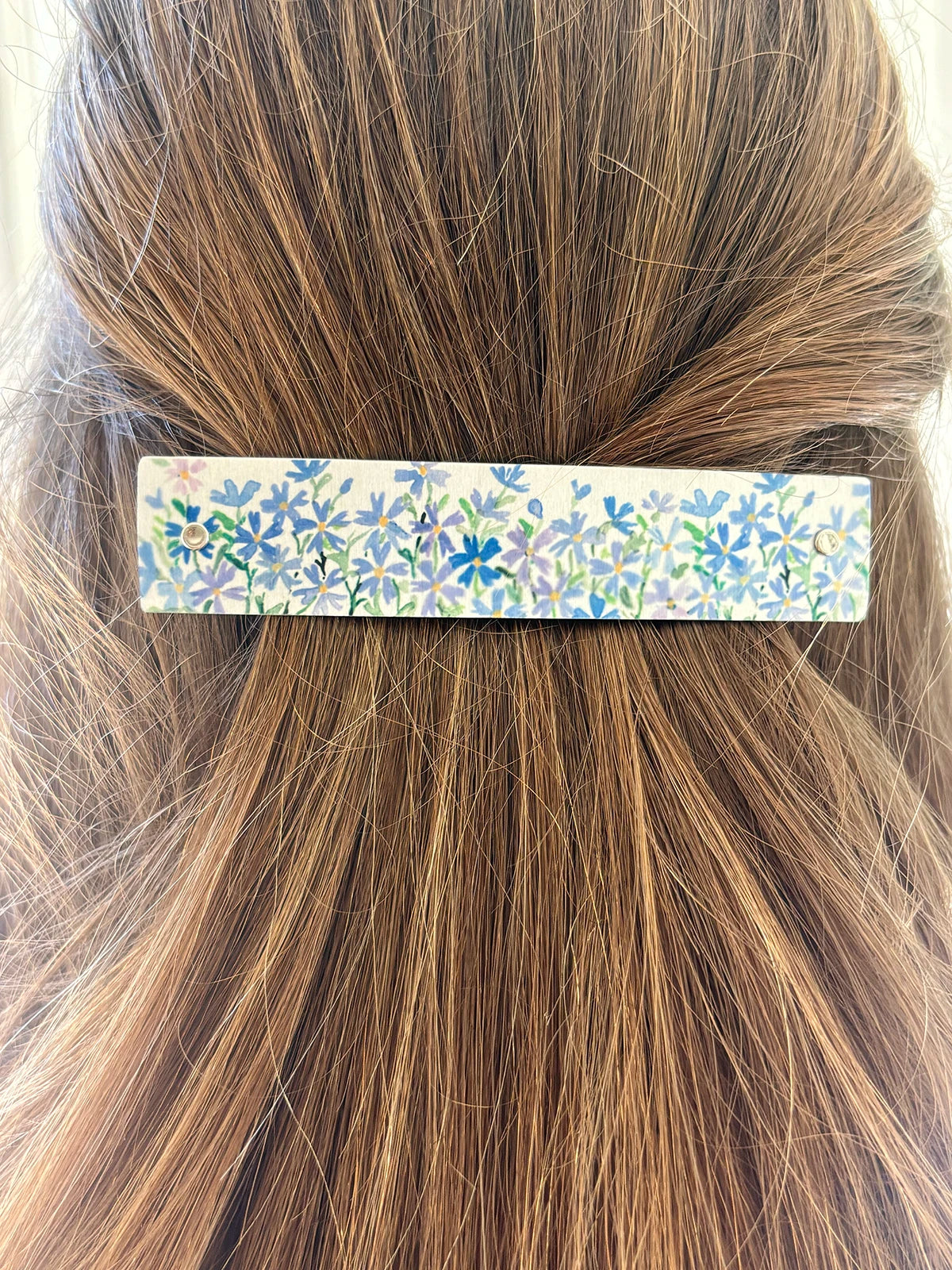 A close-up of a handmade boutique floral hair clip holding back a section of light brown hair. The rectangular hair accessory features a delicate pattern of small blue and lavender flowers with green stems on a silver metal background.