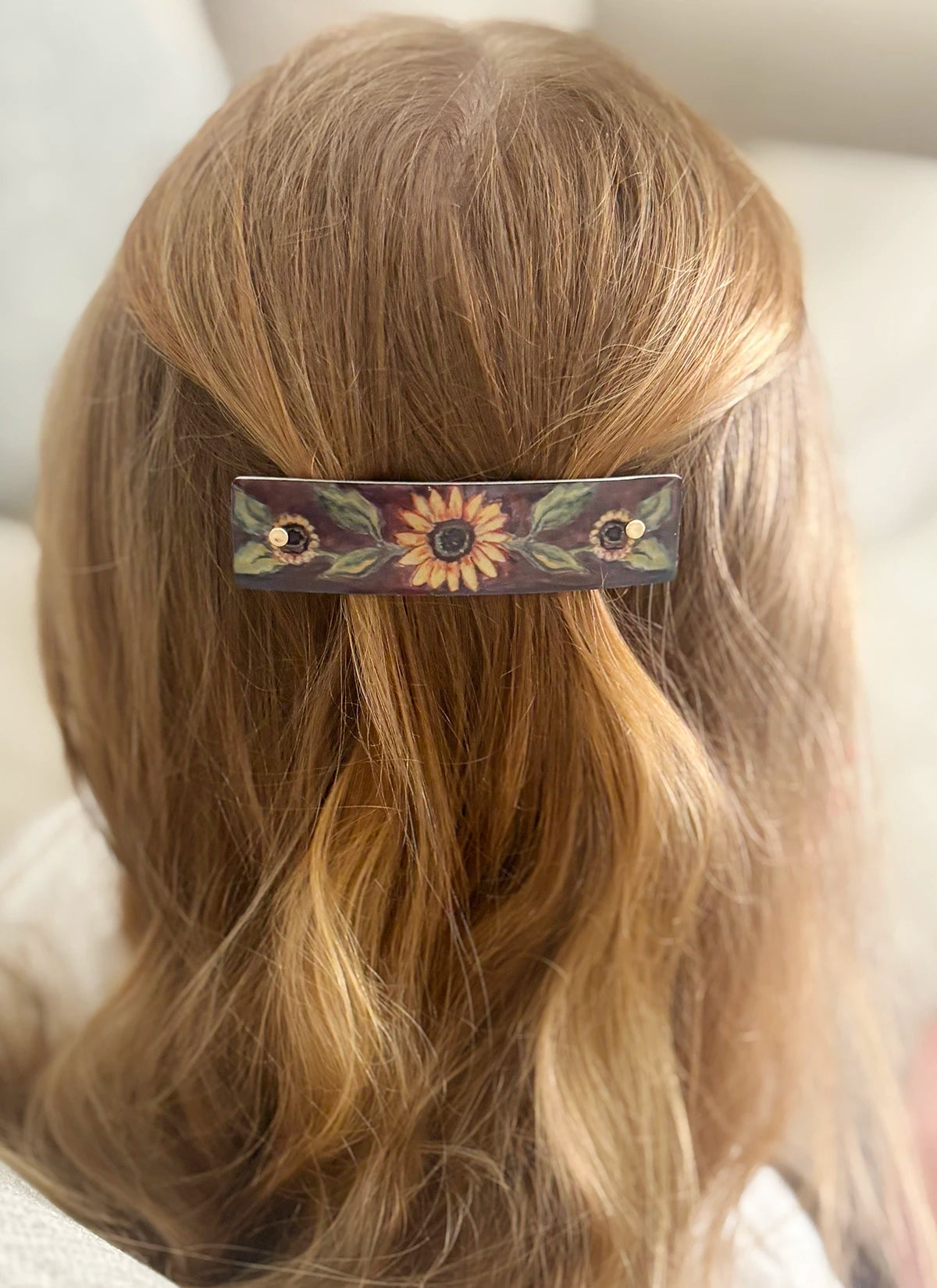A close-up back view of a person with long blond hair wearing a decorative hair clip. The rectangular clip has a dark background with a central sunflower motif and small floral details on both sides.