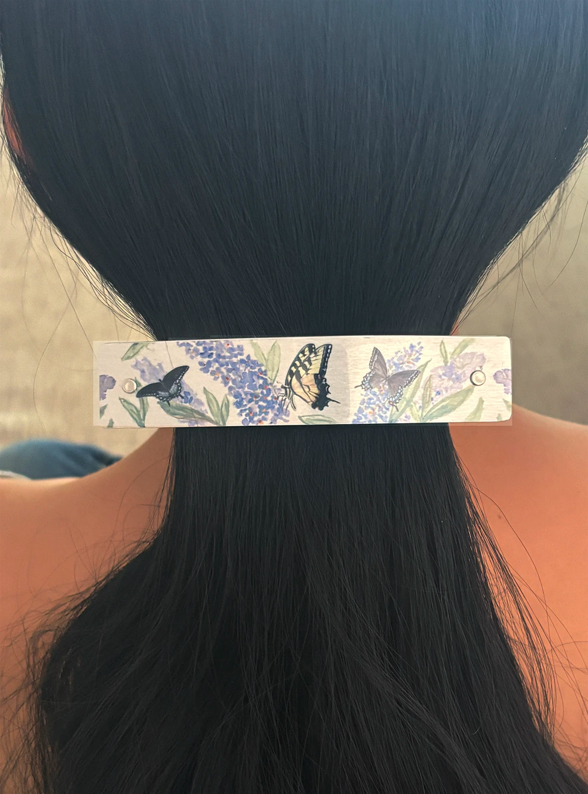 A close-up of a person's dark hair secured in a low ponytail, adorned with a long rectangular hair clip decorated with purple flowers and butterflies on a silver background.