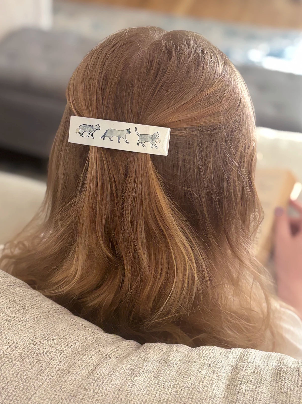 A close-up lifestyle shot of a woman's blonde hair from behind, secured with a silver rectangular hair clip decorated with a row of gray cat illustrations.