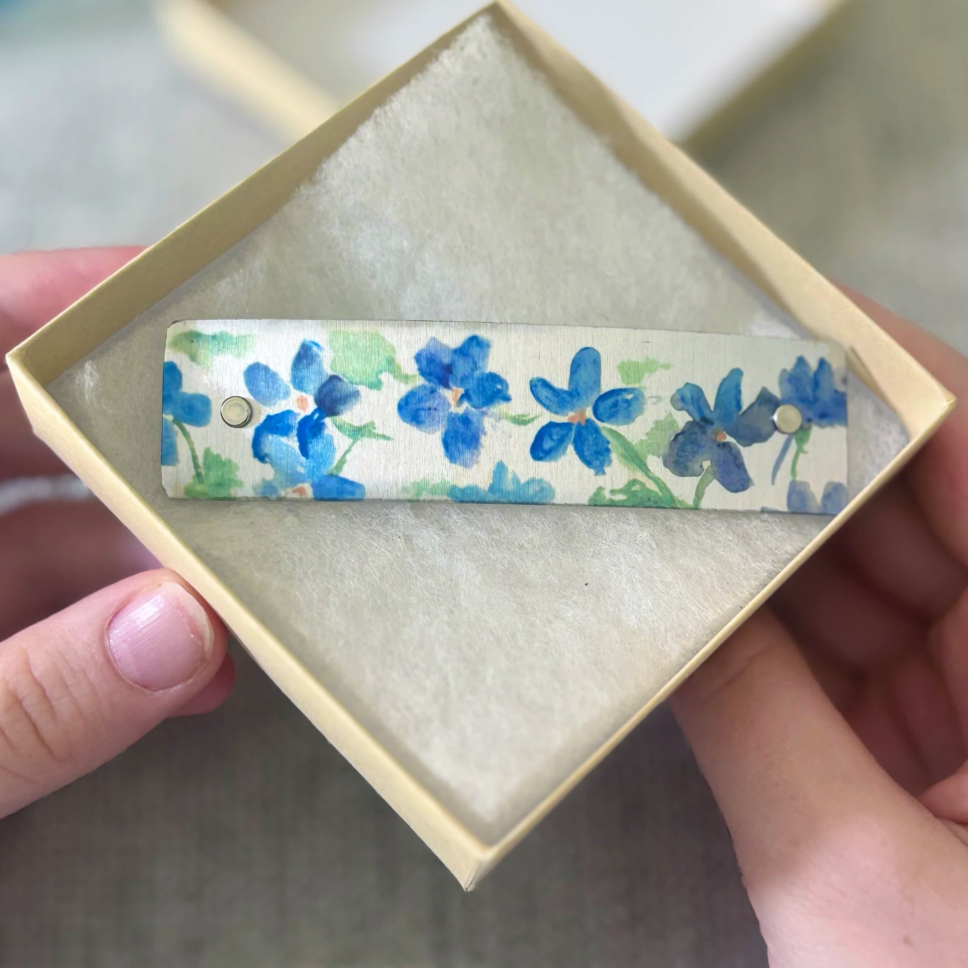 A close-up of a hand holding an open cream-colored jewelry box containing a rectangular barrette with a blue floral pattern.