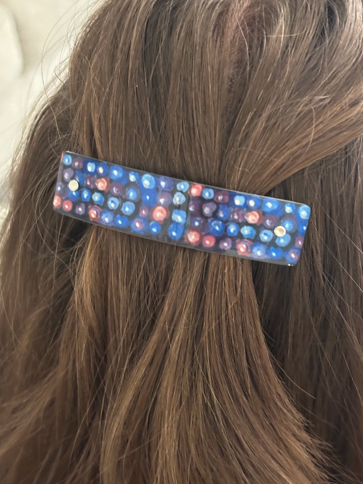 A close-up of a hair clip held in brown hair. The hair clip is rectangular and decorated with small blue, purple, and pink circles inspired by blackberries..
