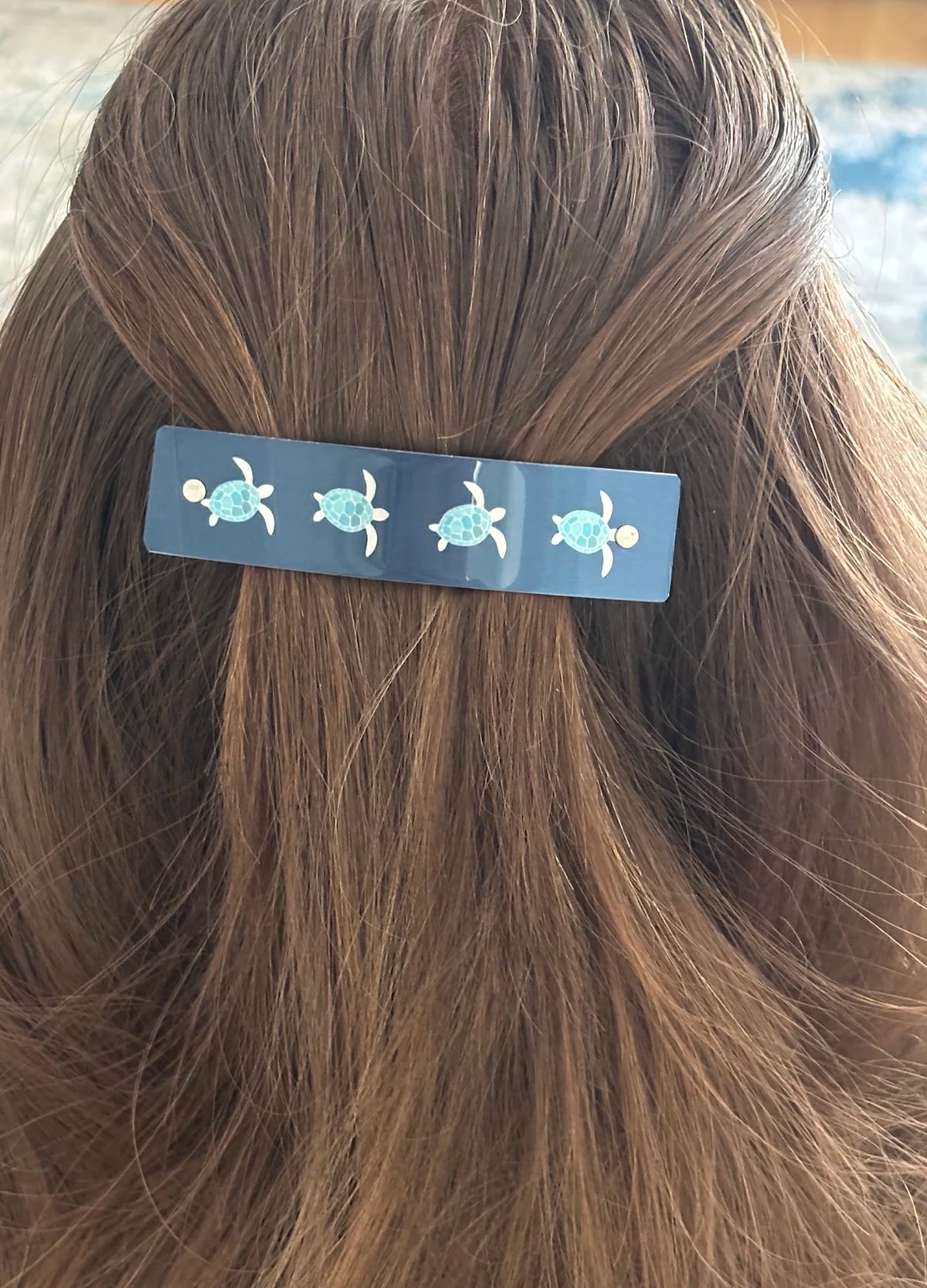 A close-up of a person's brown hair secured with a dark blue rectangular hair clip decorated with four small sea turtle illustrations.