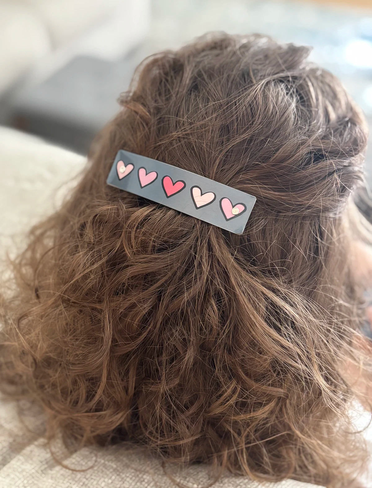 A close-up of a person's brown curly hair secured with a gray hair clip decorated with pink hearts.