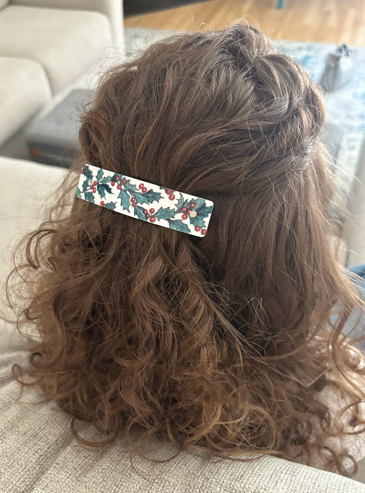 A close-up back view of a person with long, wavy brown hair wearing a rectangular hair clip decorated with a holly and berries pattern, sitting on a light-colored couch.