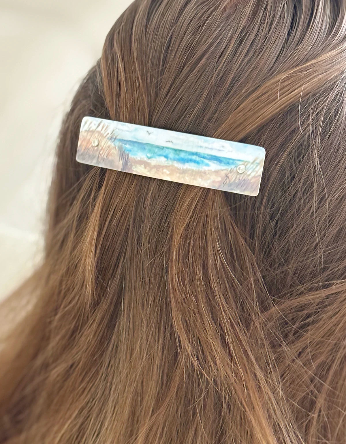 A close-up of a person's brown hair holding a rectangular handmade hair clip with a blue and white watercolor beach scene design.