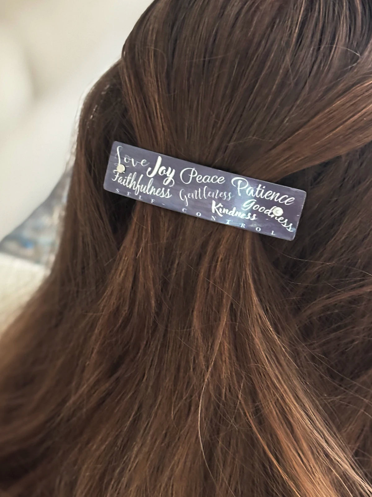 A close-up of a person's brown hair holding a small rectangular hair clip with silver script words including 'Love', 'Joy', 'Peace', 'Patience', 'faithfulness', 'gentleness', 'goodness', and 'kindness'.