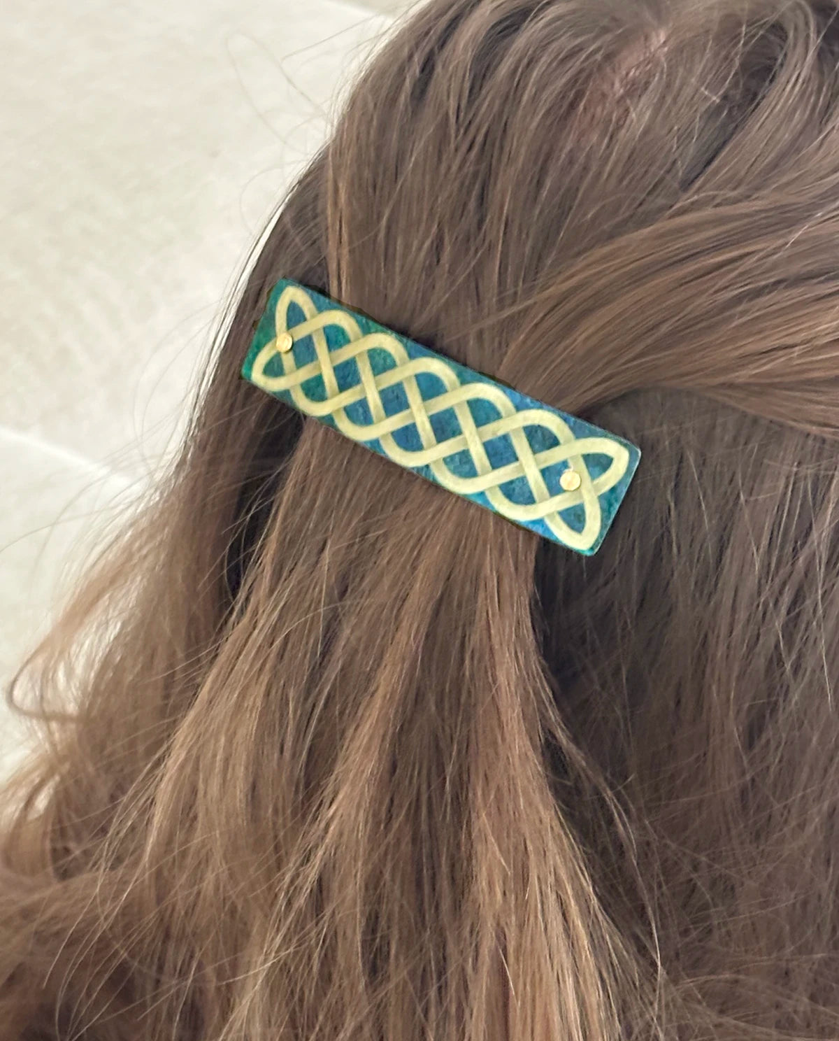 A close-up of light brown hair secured with a rectangular hair clip featuring a gold-colored Celtic knot pattern over a green background, by Robbin V. Davis Studio. Handmade in North Carolina.