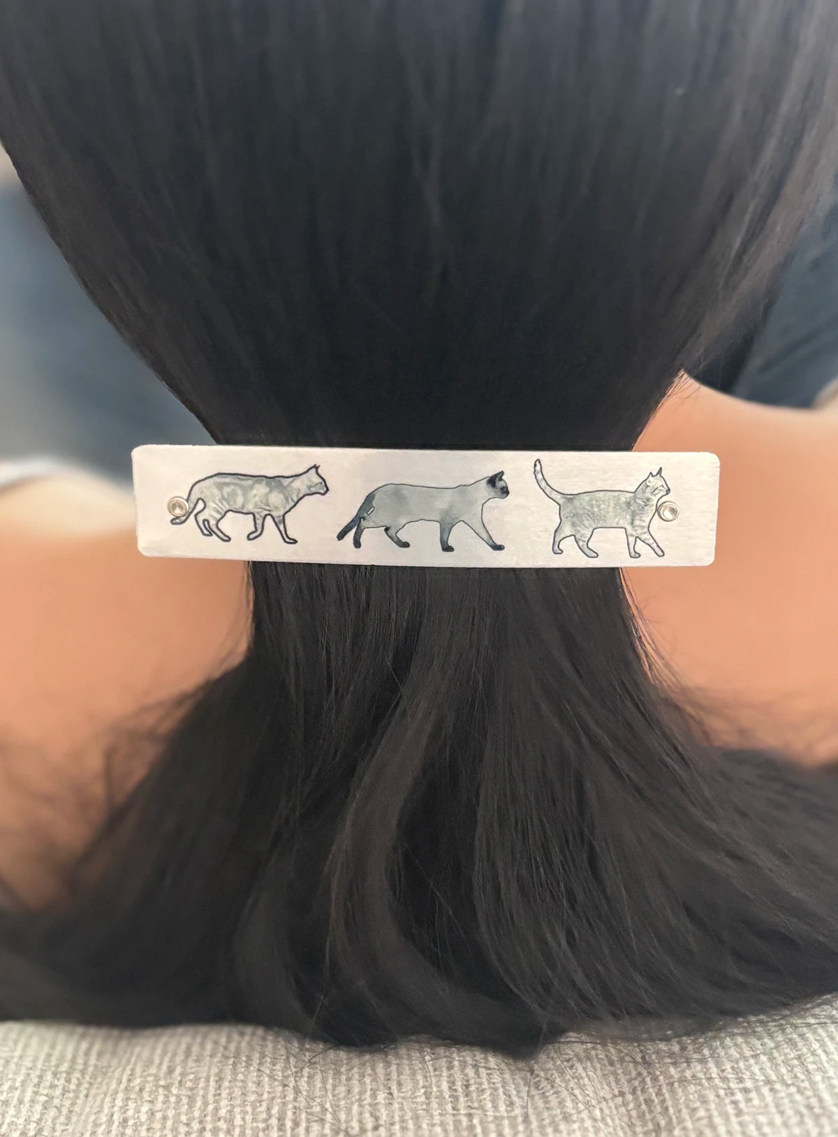 A close-up of a person's dark hair in a ponytail, secured with a silver rectangular hair clip decorated with three gray cat illustrations.