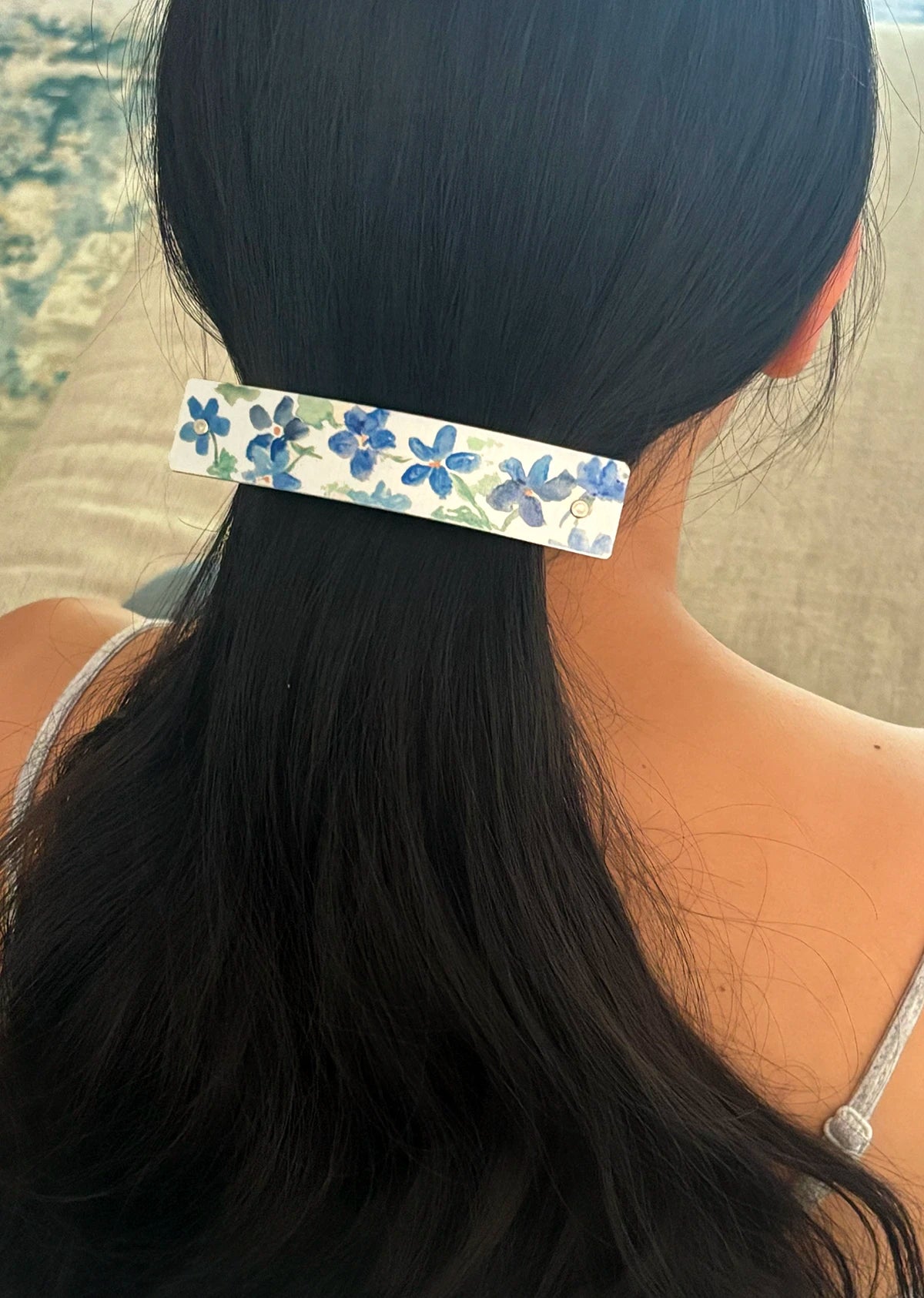 A close-up lifestyle shot of a woman's dark hair secured in a ponytail with a silver metal hair clip decorated with an artistic watercolor of blue flowers and green leaves.