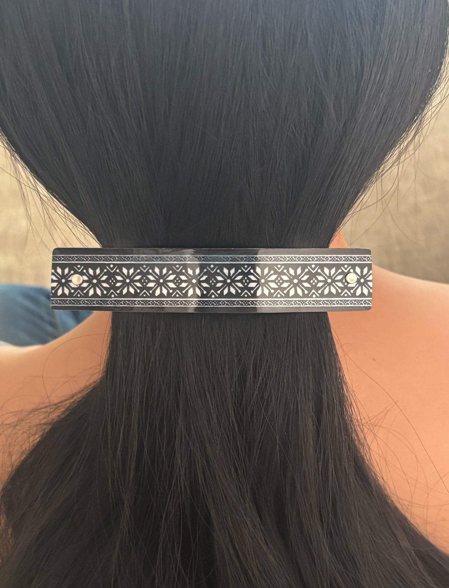 A close-up of a person's dark hair clipped back in a ponytail, wearing a rectangular black hair barrette with an intricate white geometric floral pattern.