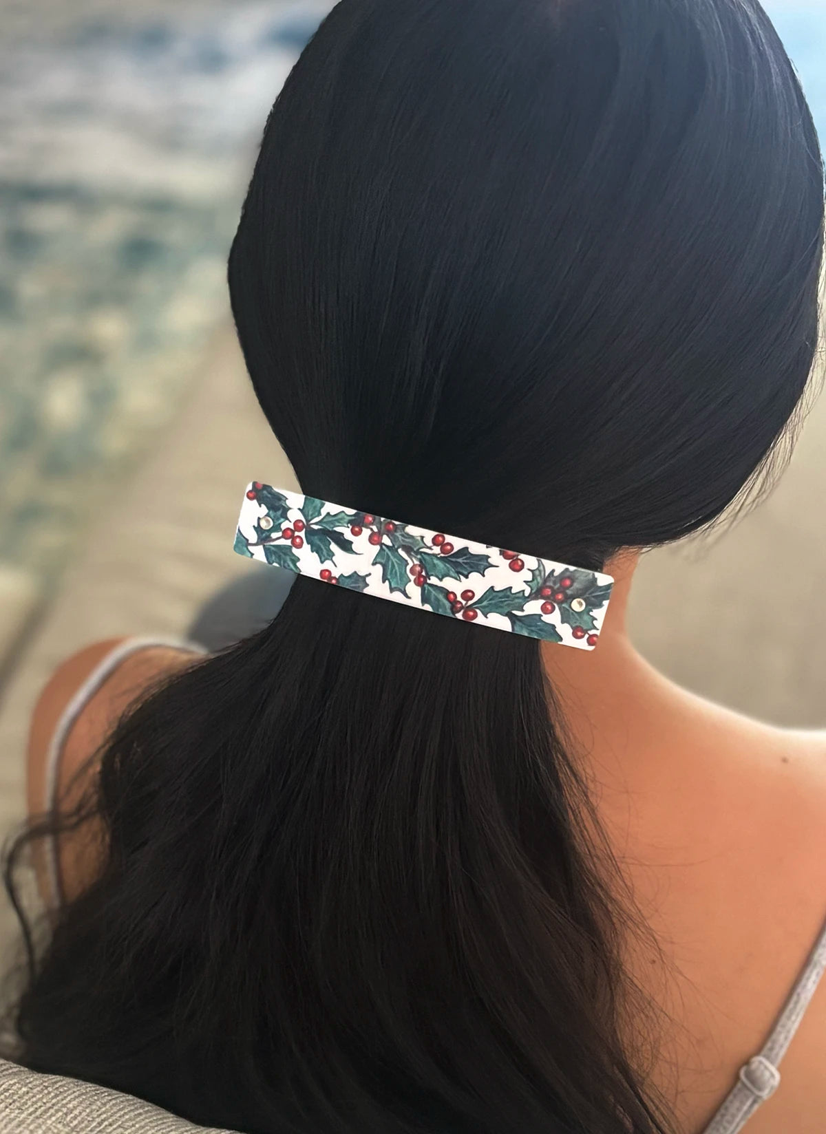 A close-up back view of a person with long black hair secured in a low ponytail, wearing a white hair clip decorated with a green holly and red berry pattern.