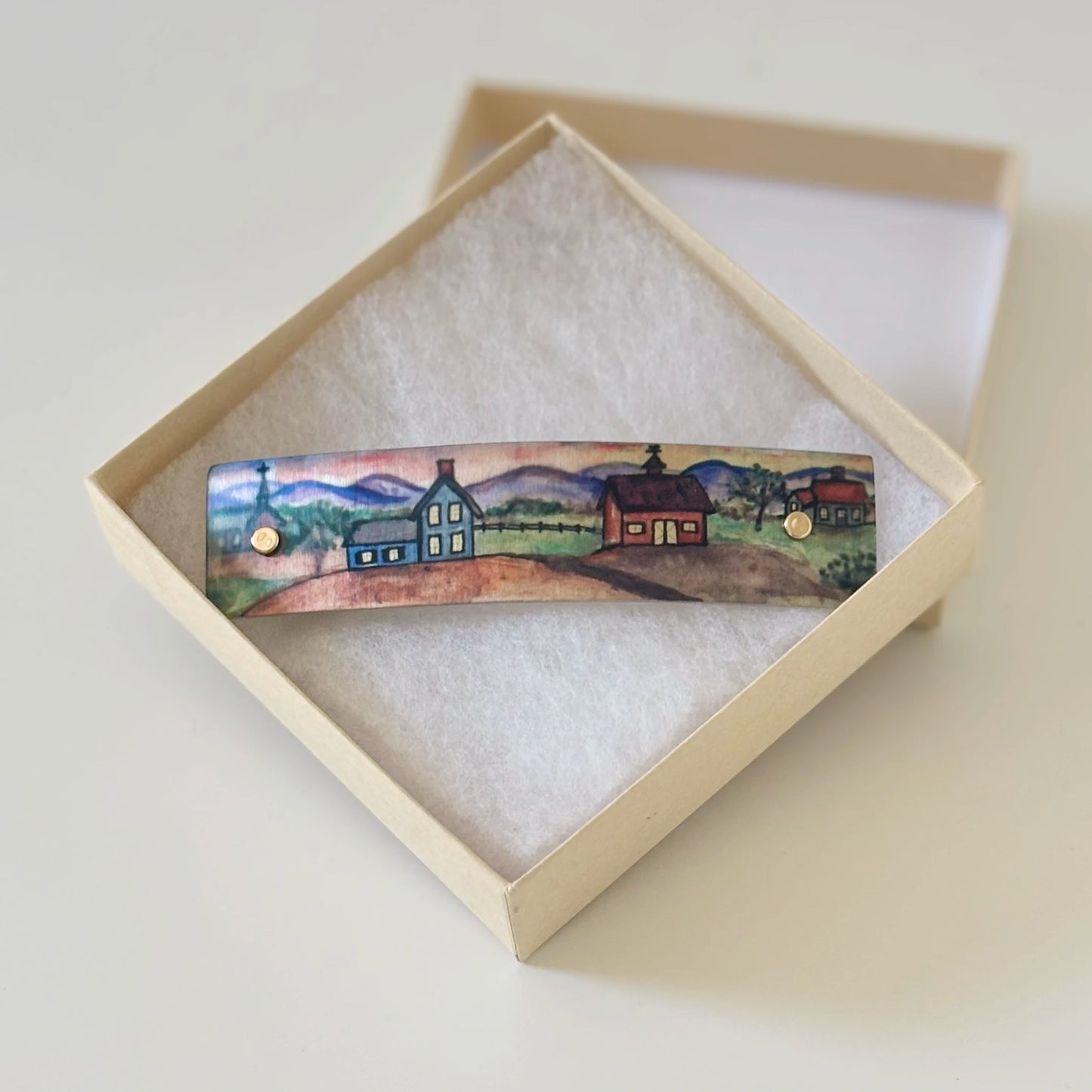 A hand-painted metal boutique hair clip  featuring a small rural landscape with houses and mountains, displayed inside an open cream-colored presentation box with white padding.