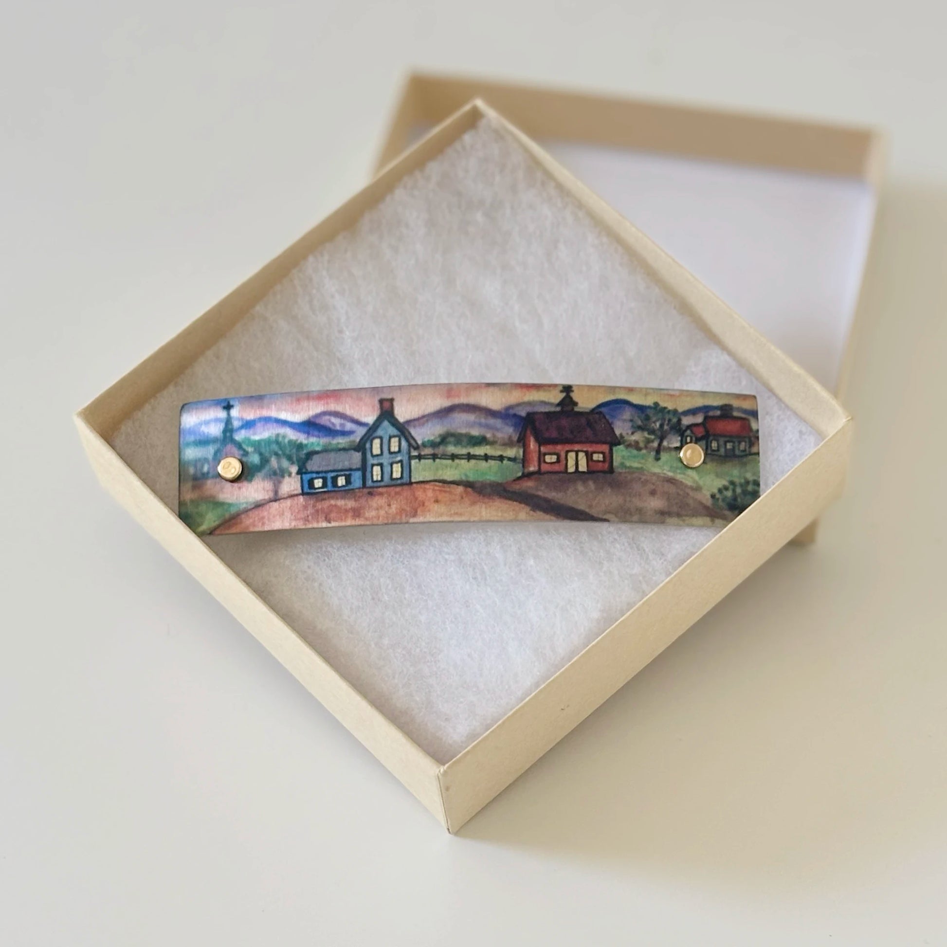 A hand-painted metal boutique hair clip  featuring a small rural landscape with houses and mountains, displayed inside an open cream-colored presentation box with white padding.