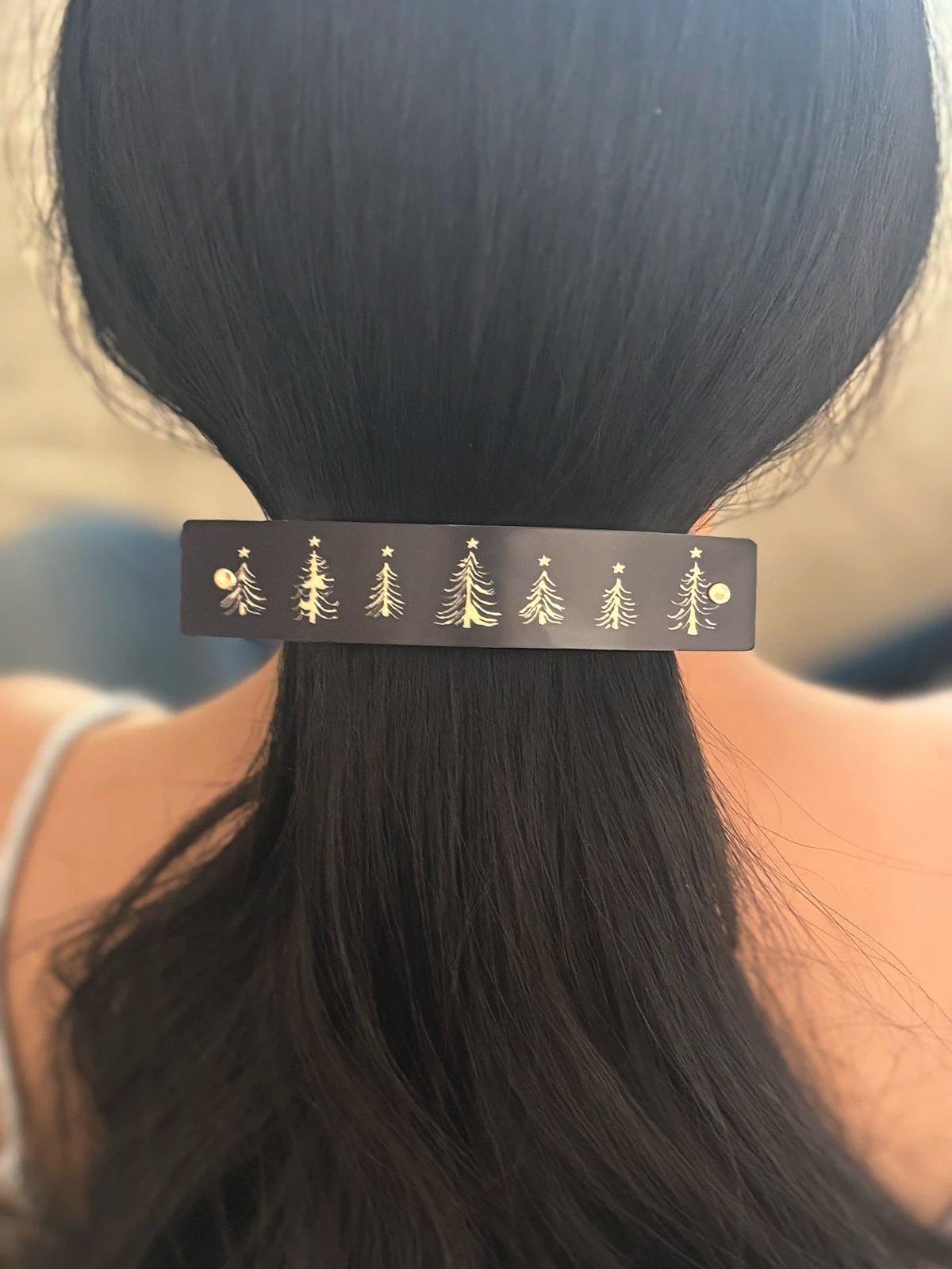 A close-up of a person's dark hair secured in a low ponytail with a black rectangular hair clip decorated with small gold Christmas tree and star designs.