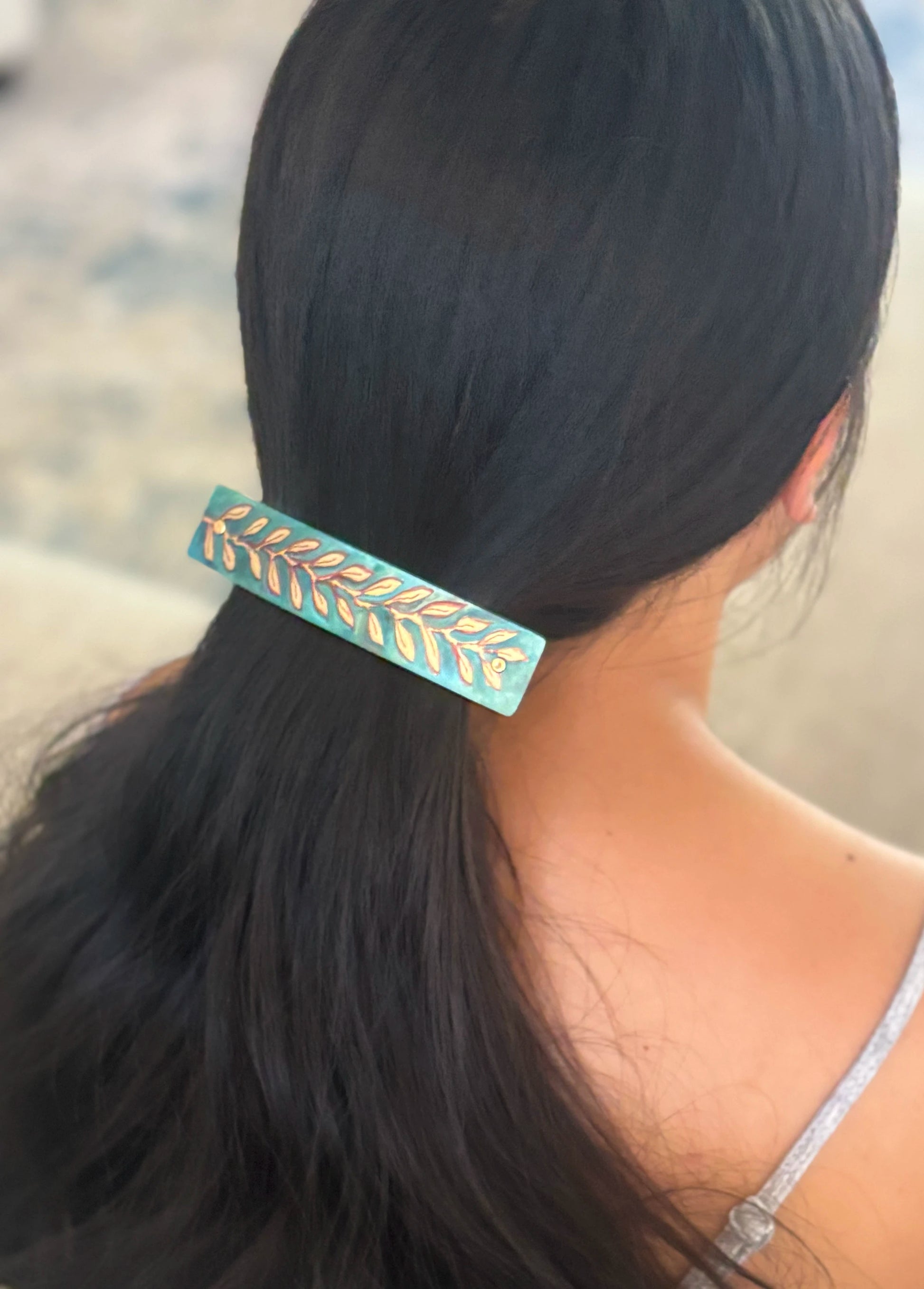 A close-up lifestyle shot of a woman with long black hair wearing a rectangular hair clip. The clip is light blue with a gold leaf pattern, holding her hair in a low ponytail.