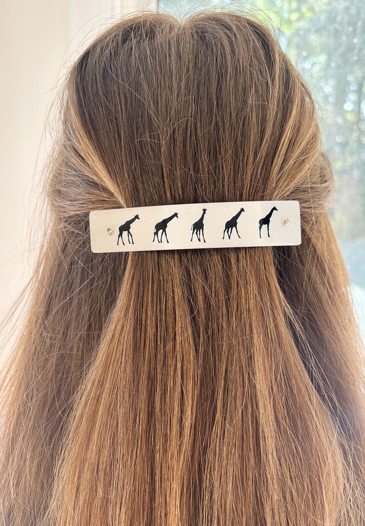 A close-up of a person's long, light brown hair secured with a white rectangular hair clip decorated with black giraffe silhouettes, set against a softly blurred indoor background.