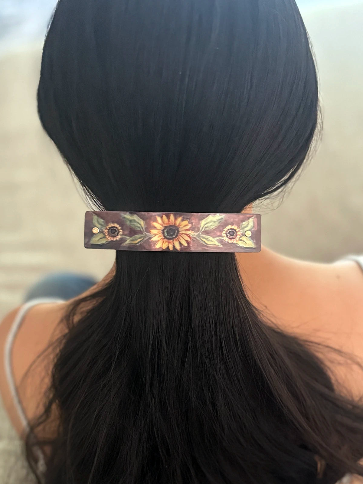 A close-up back view of a person with long black hair wearing a decorative hair clip. The clip is rectangular and features a floral sunflower design in gold, brown, and green tones against a dark background.