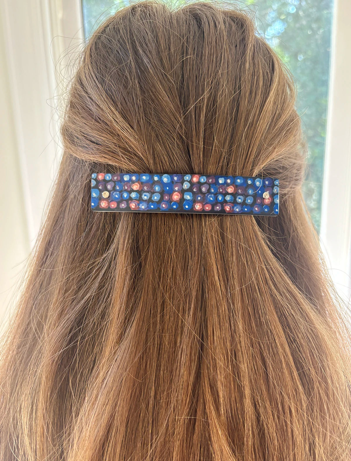 A close-up back view of a person with long light brown hair wearing a hair clip adorned with a blackberry design in blues, blacks, pinks, and purples.