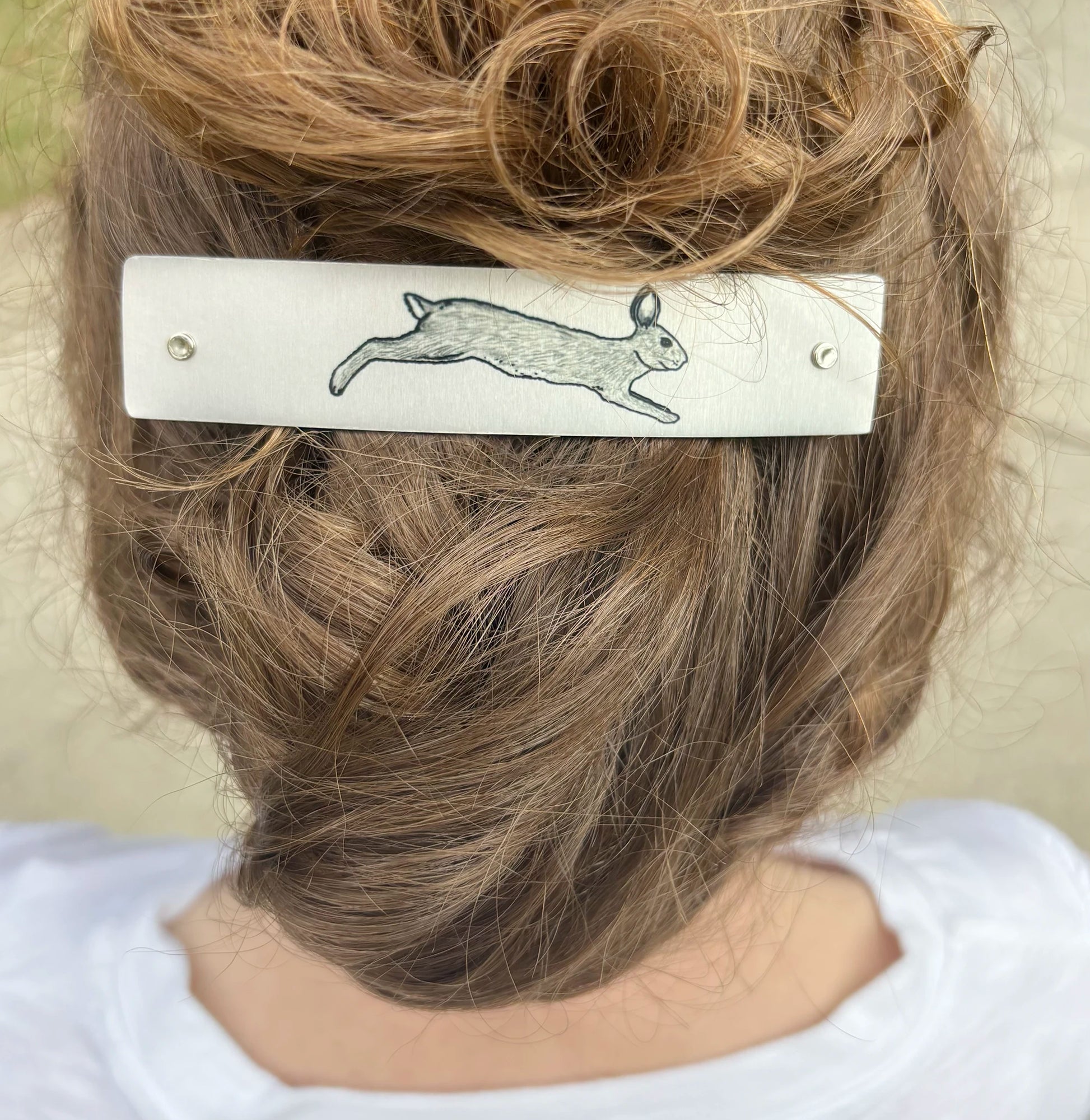 A close-up back view of a person wearing a silver metal hair clip with a hand drawn rabbit design, holding up light brown hair.