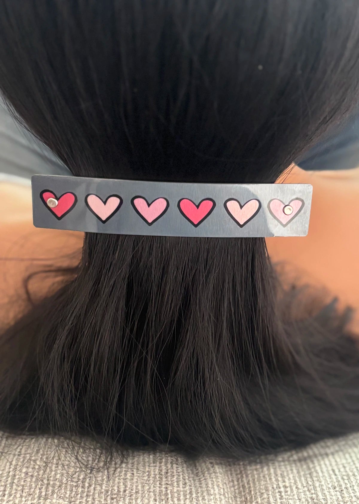 A close-up of a person's dark hair secured with a gray hair clip decorated with six pink hearts.