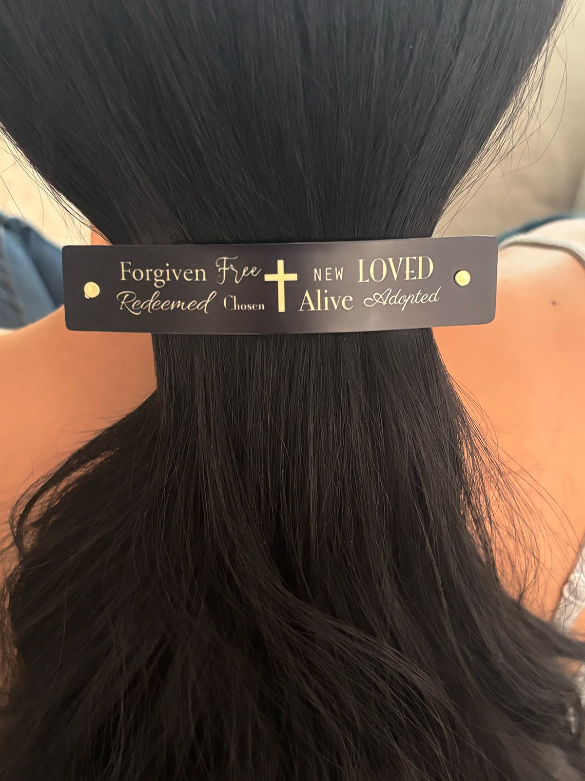 A close-up of a person's dark hair secured with a black hair clip featuring gold text and a gold cross. The text on the clip reads 'Forgiven Free Redeemed Chosen NEW LOVED Alive Adopted'.