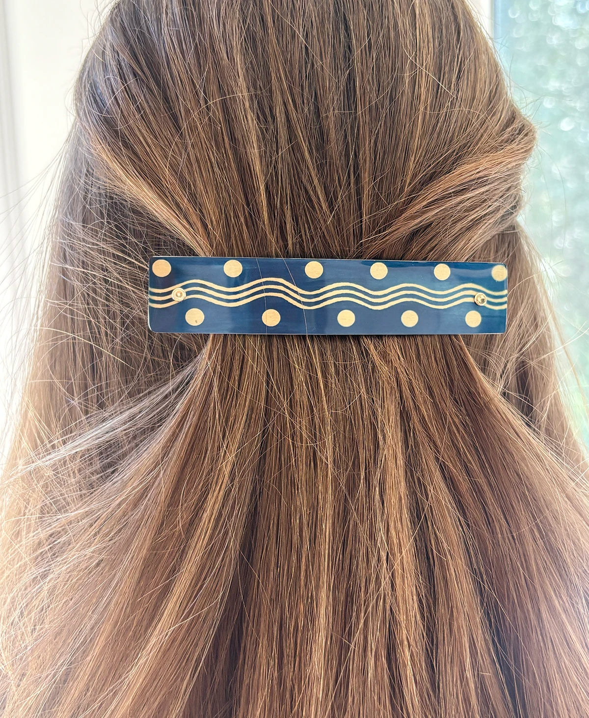 A close-up of a person's brown hair secured with a navy blue hair clip decorated with gold polka dots and wavy gold lines.
