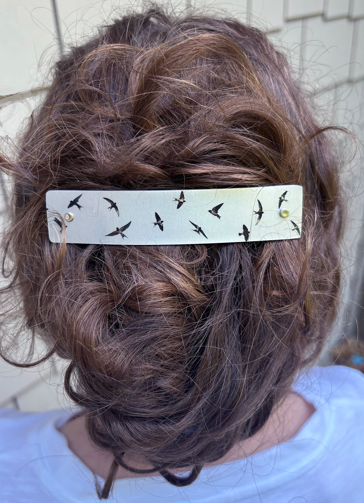 A close-up back view of a person wearing a silver hair clip adorned with small black bird silhouettes, holding up a styled brown updo.