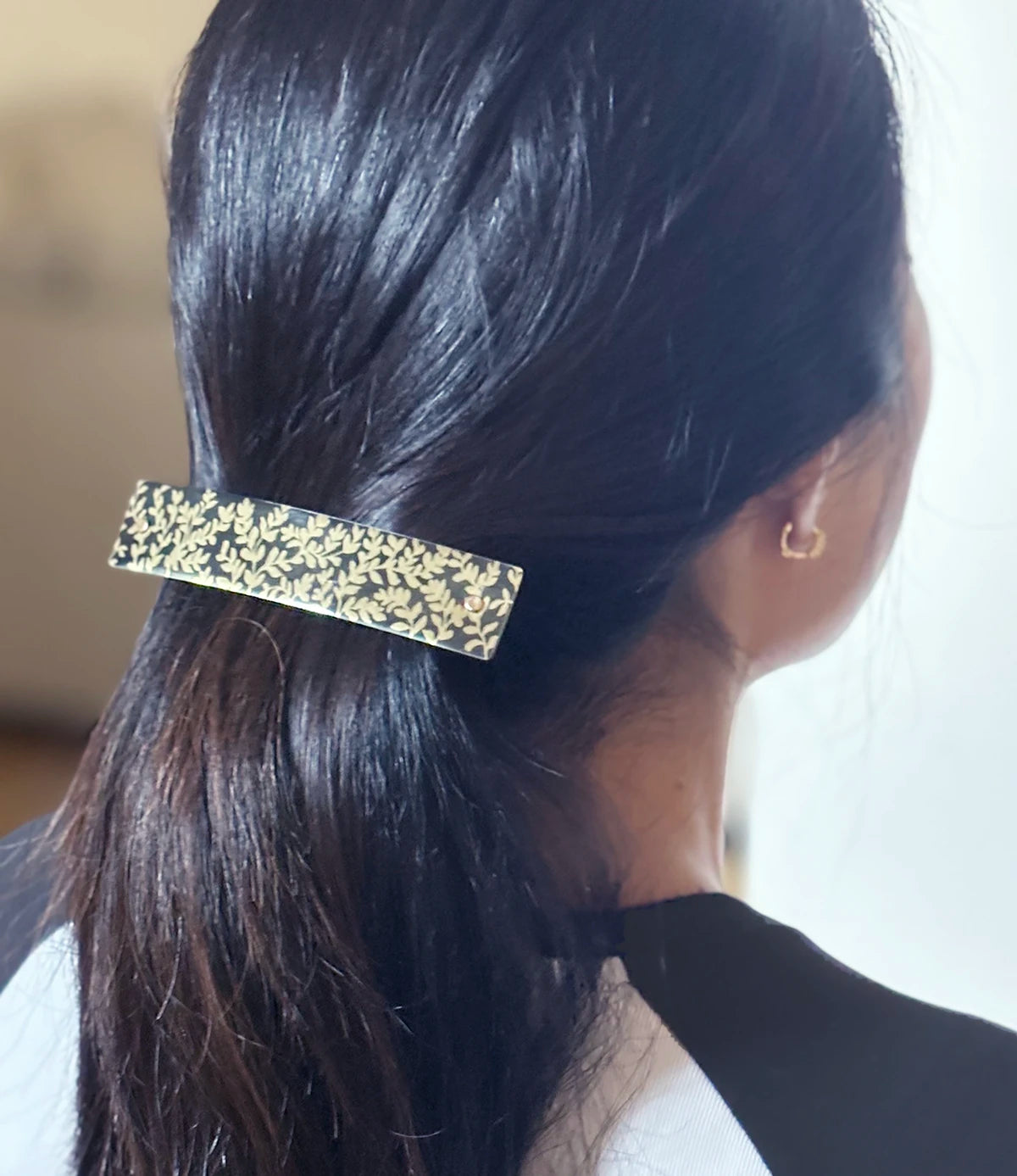 A close-up back view of a person with dark hair secured with a rectangular hair clip featuring a gold-toned leaf pattern, wearing a small gold hoop earring.