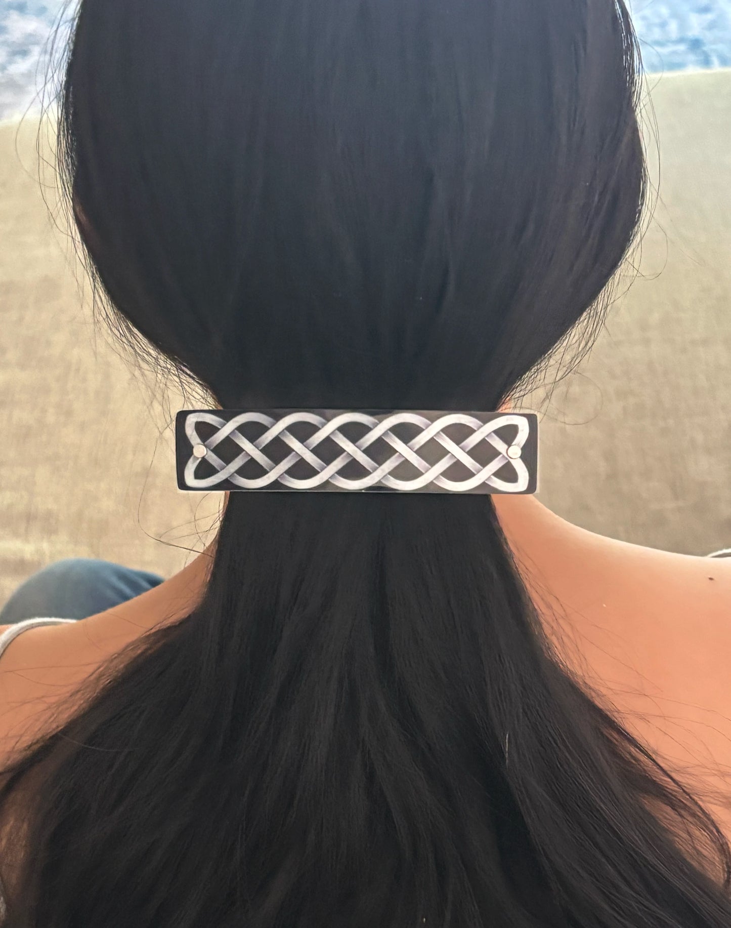 A close-up back view of a person with long black hair gathered in a ponytail, wearing a rectangular black hair clip with a silver Celtic knot pattern.