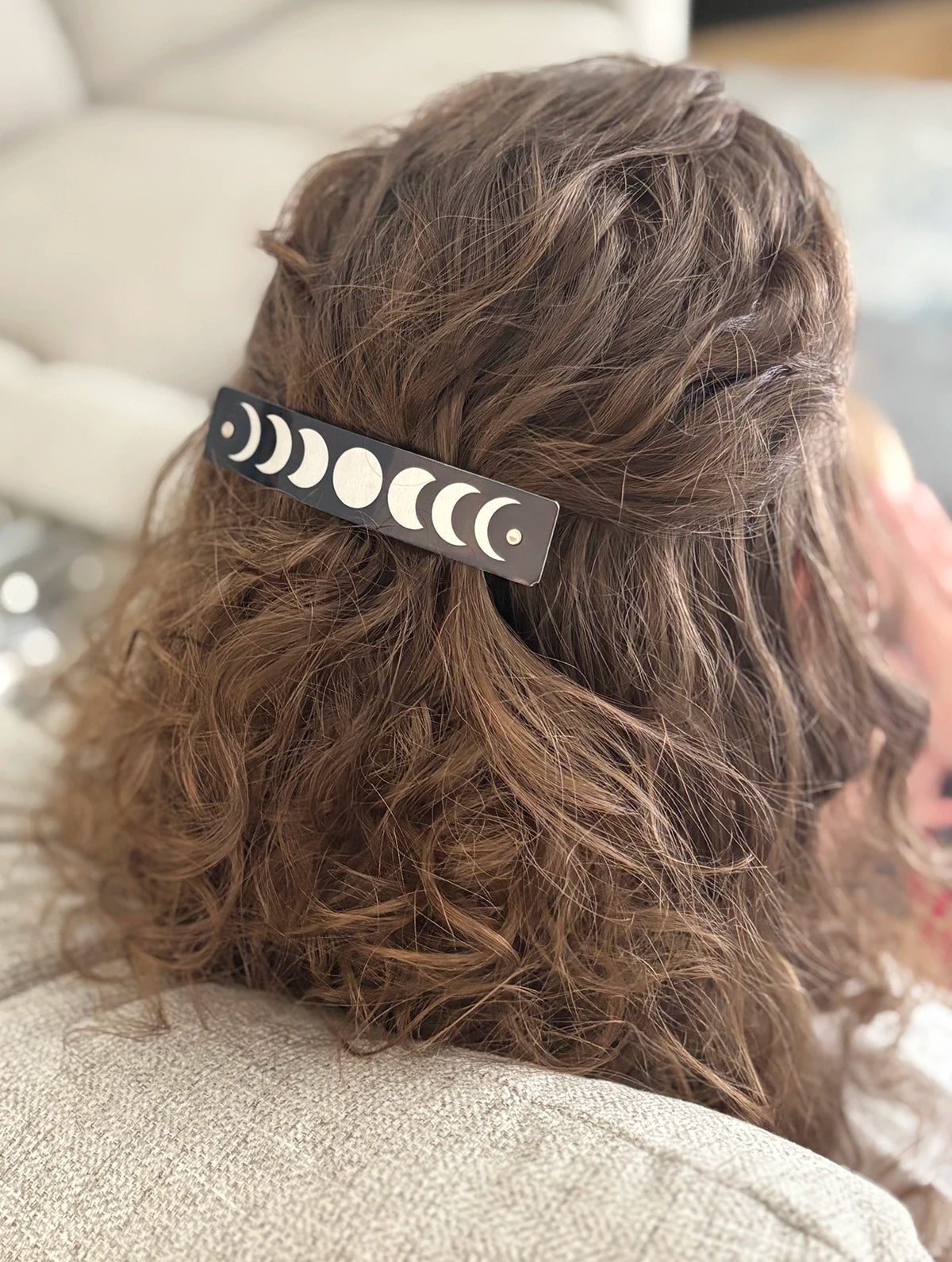 A close-up lifestyle shot of a person's brown wavy hair secured with a black barrette hair clip featuring a silver moon phase design.