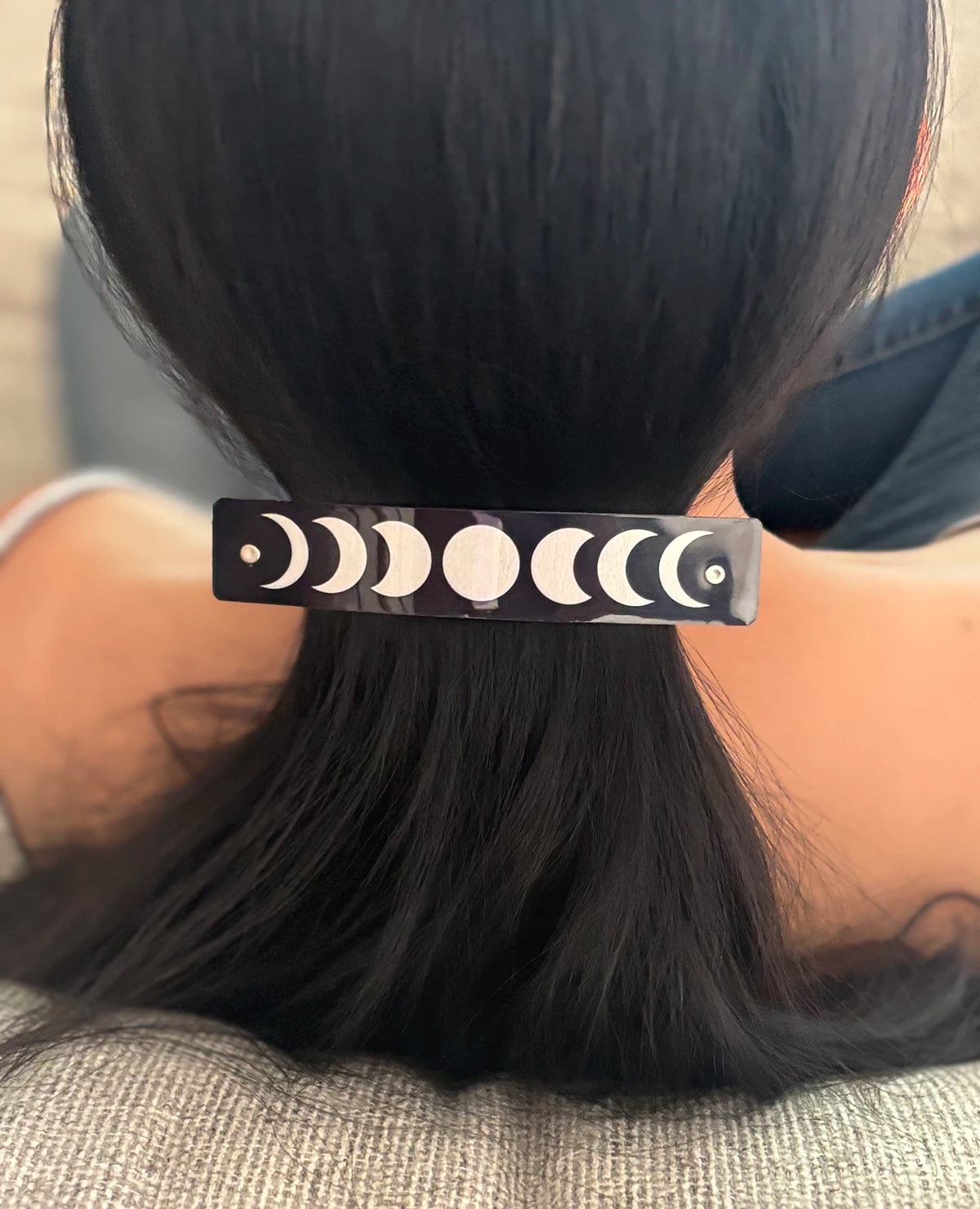A close-up lifestyle shot of a person's dark hair secured with a rectangular black hair clip decorated with a silver moon phase pattern.