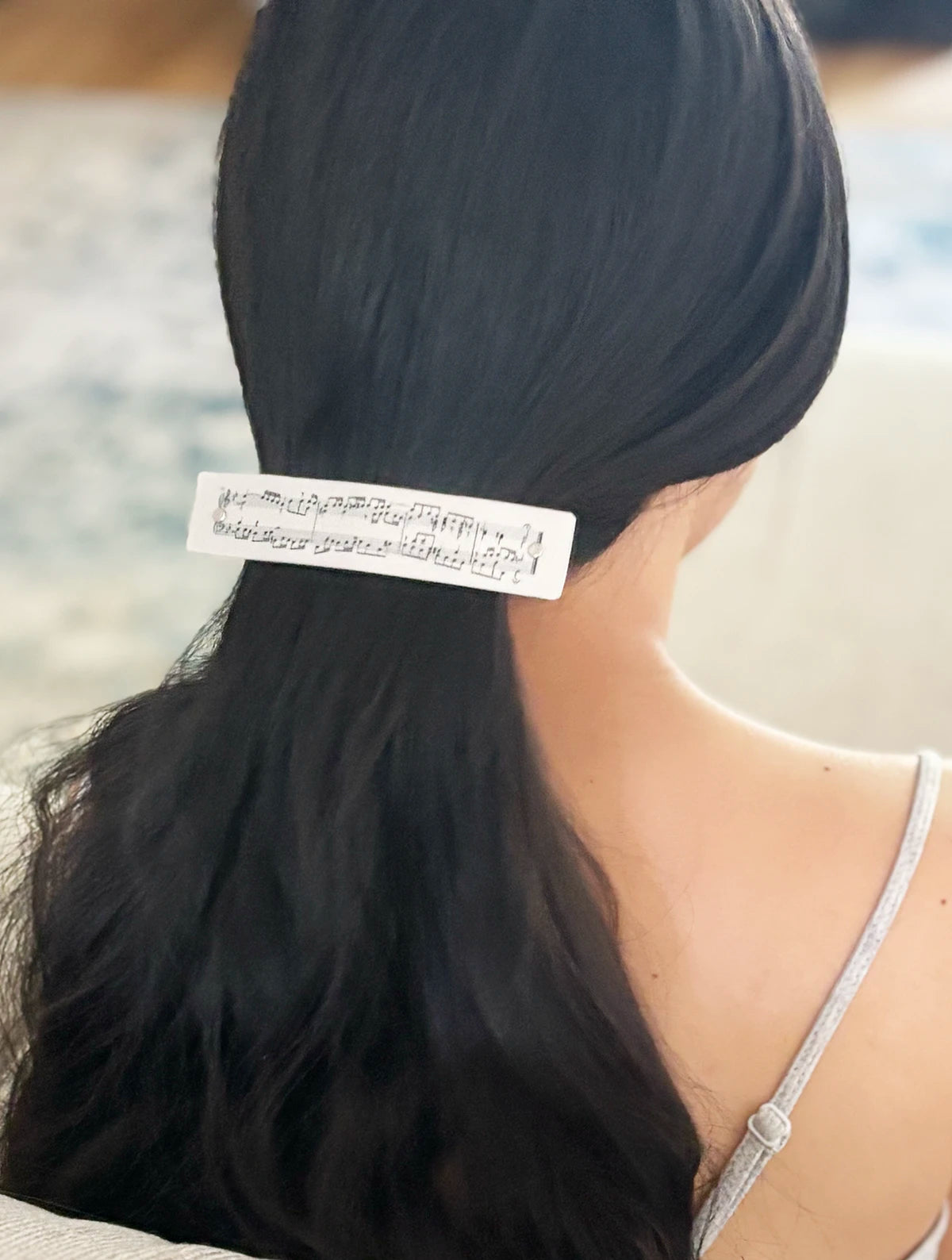 A close-up back view of a person with long, straight black hair wearing a wsilver boutique hair clip decorated with black musical notes, wearing a light gray spaghetti-strap top.