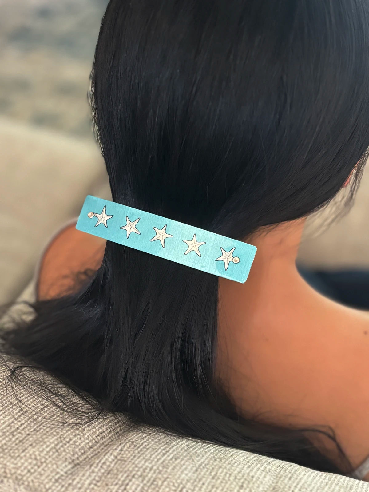 A light blue rectangular hair clip with five silver starfish designs, shown worn on a person's dark hair.