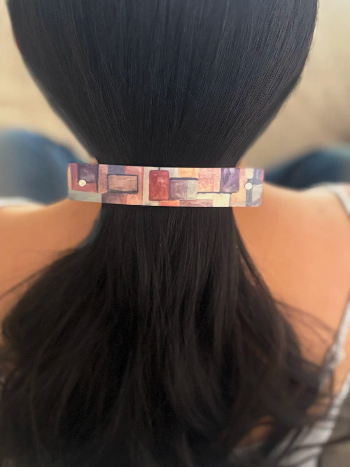 A close-up back view of a person with long dark hair wearing a slim rectangular French barrette with a multicolor geometric pattern.