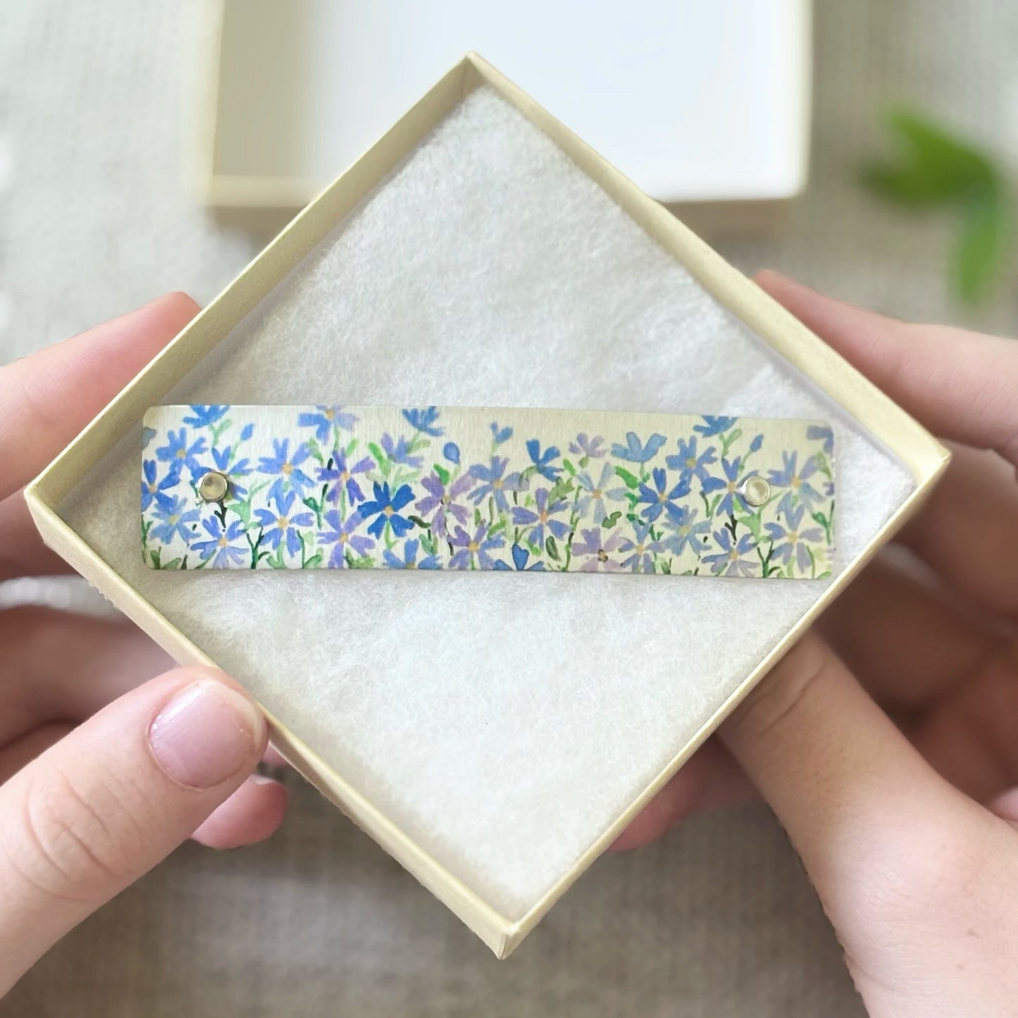 A floral  boutique hair clip with blue, purple, and green flowers and leaves on a silver metal background, held inside a square beige gift box by two hands.