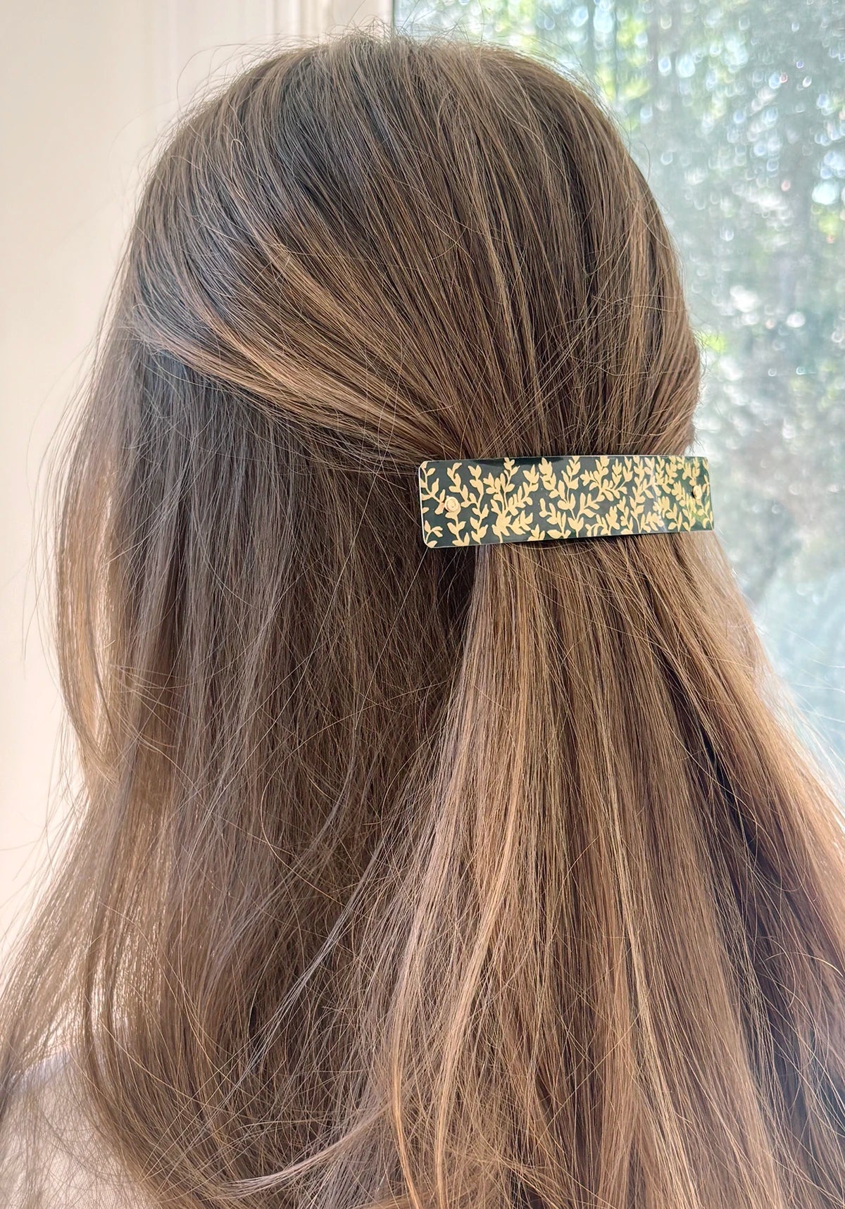 A close-up of a person's brown hair secured with a slim rectangular hair clip featuring a gray and gold leaf pattern, set against a softly blurred window background.