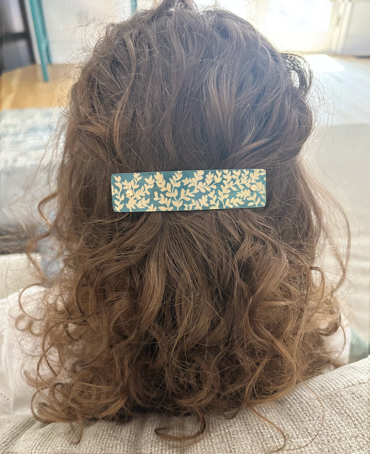 A close-up back view of a person with long, wavy brown hair wearing a rectangular hair clip. The clip has a teal green background with a delicate gold leaf pattern.