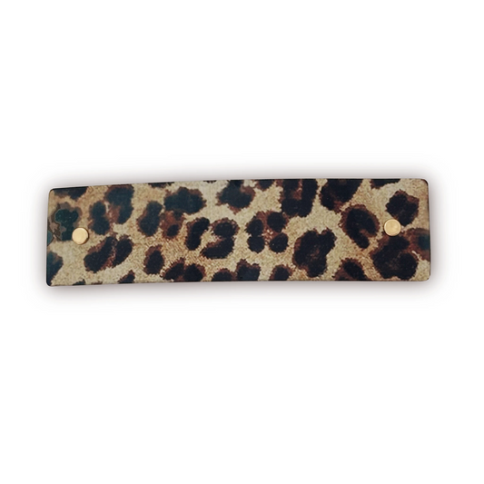 A rectangular metal hair clip accessory with a leopard print pattern in beige, brown, and black, featuring two small gold-tone metal rivets on the left and right edges, shown on a plain white background.