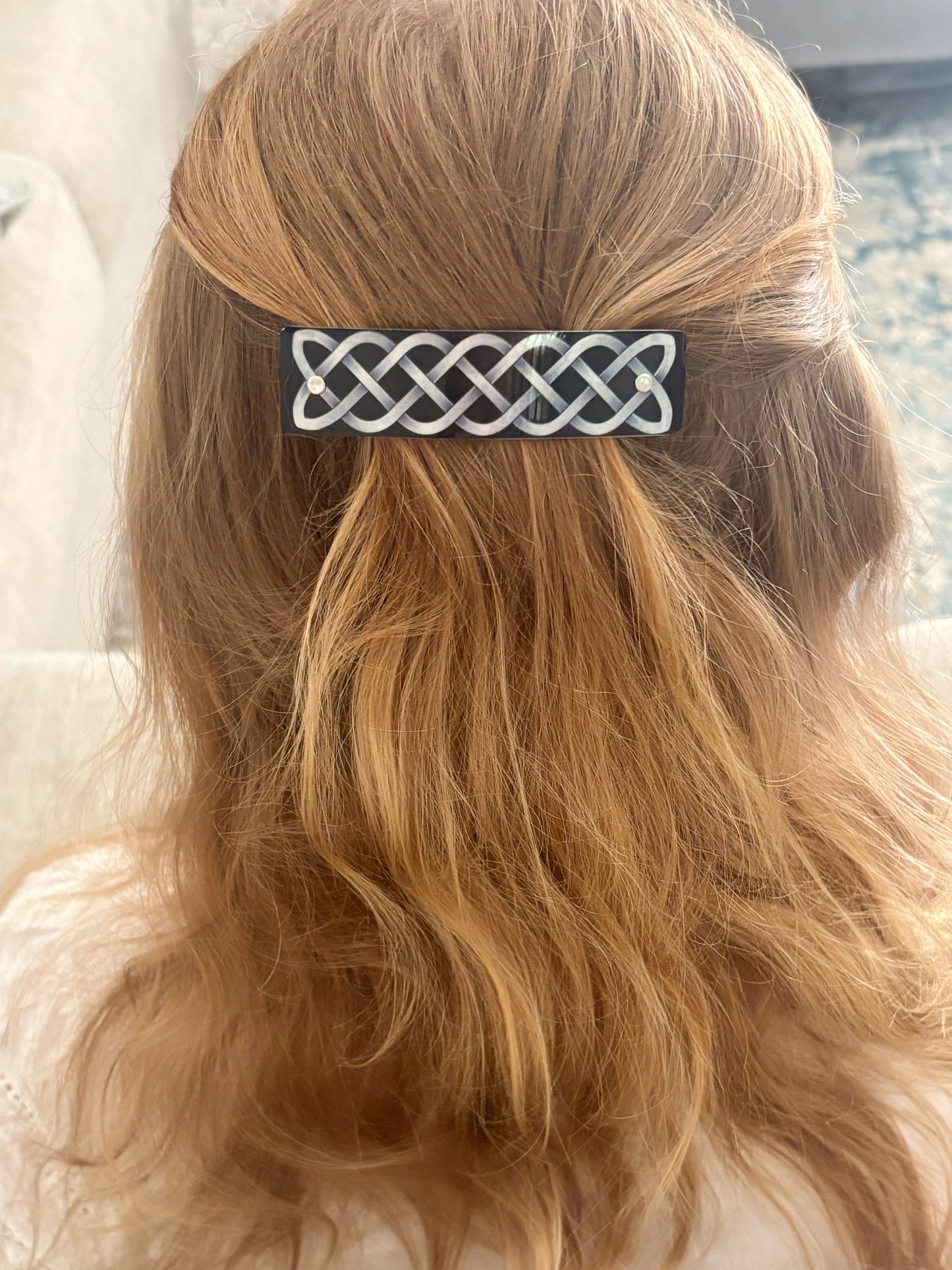 A close-up back view of a person with light brown hair wearing a rectangular black hair clip decorated with a silver Celtic knot pattern.
