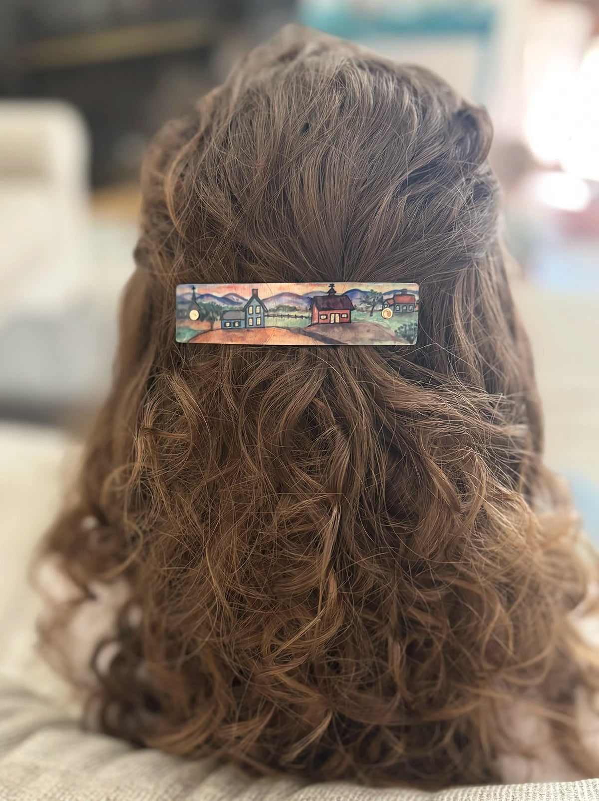 A close-up back view of a person with long, wavy brown hair wearing a small rectangular hair clip. The clip features a colorful landscape illustration with houses, trees, and a sunset sky.