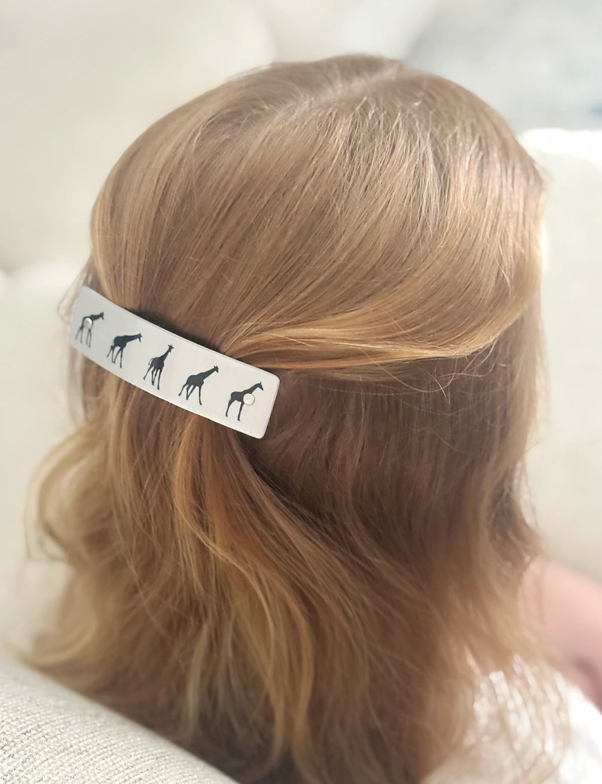 A close-up of a person's blonde hair secured with a silver hair clip decorated with black giraffe silhouettes, set against a soft, light-colored background.