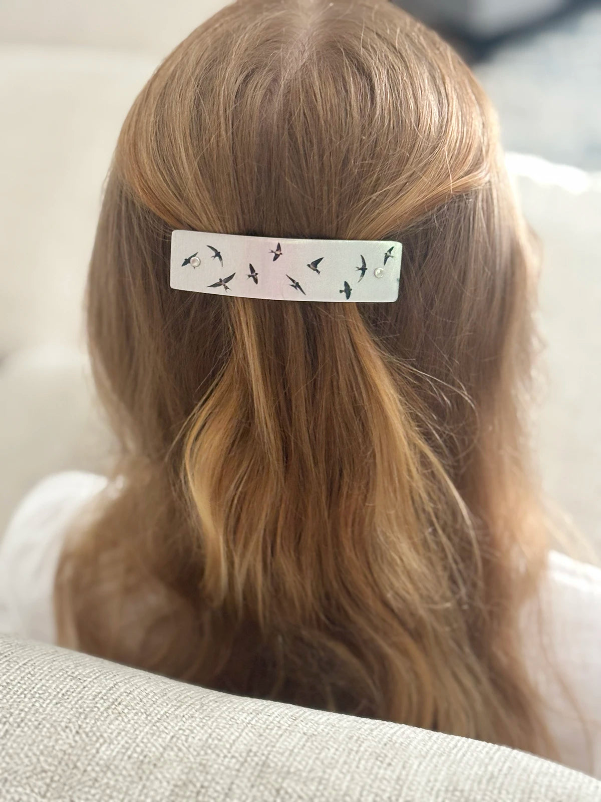 A close-up back view of a person with long strawberry blond hair wearing a silver hair clip decorated with small black flying swallow bird silhouettes.