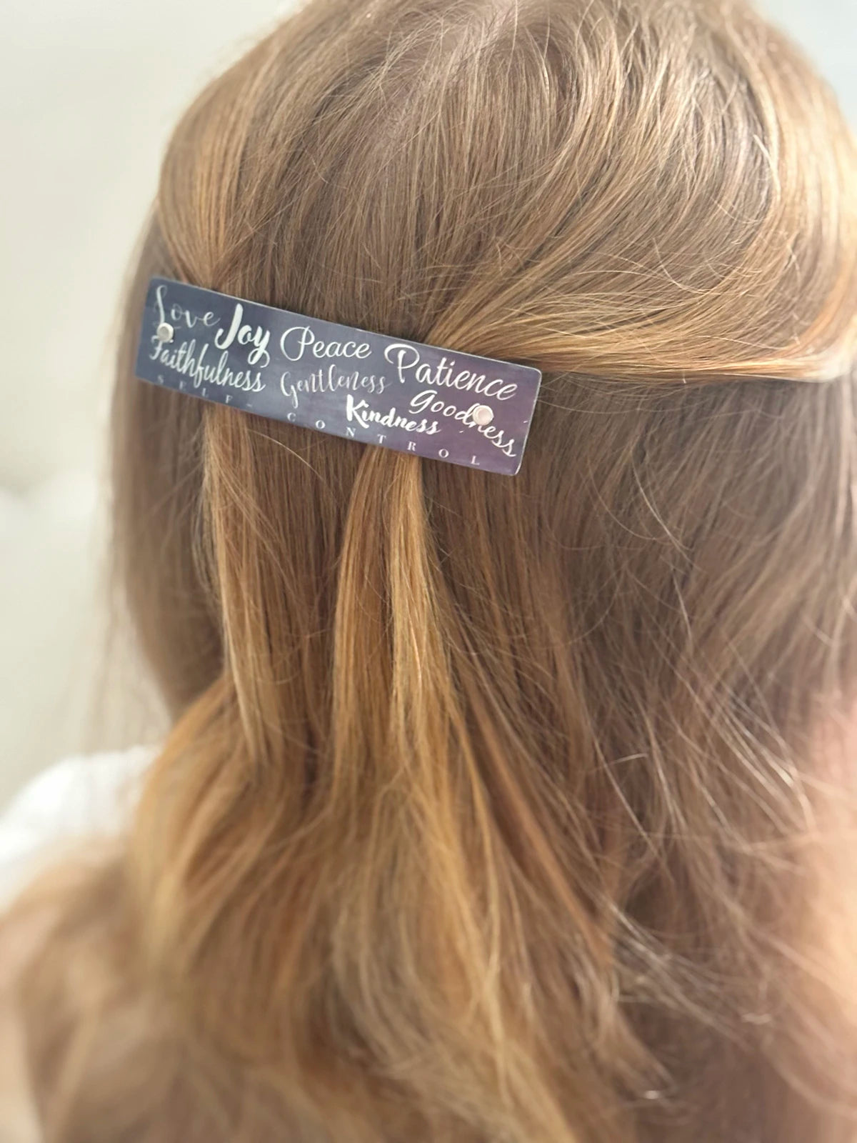A close-up of a person's strawberry blonde hair with a medium rectangular hair clip holding a section of hair. The clip is dark gray with silver cursive text listing virtues: 'Love, Joy, Peace, Patience, Faithfulness, Gentleness, Goodness, and Kindness'.