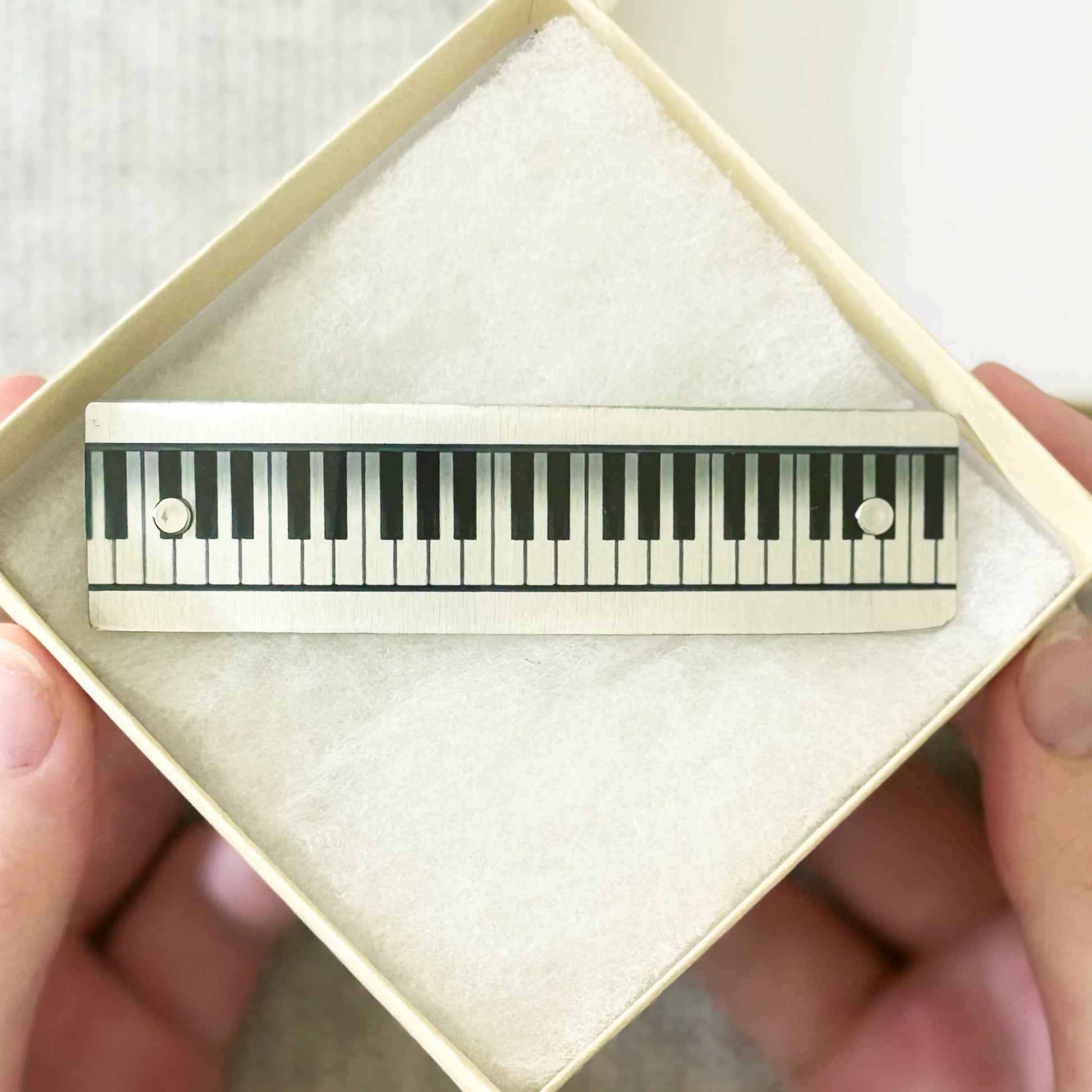 A piano-key themed hair clip with black and silver keys, held inside an open cream-colored gift box with a soft white insert. Human hands are visible holding the box.
